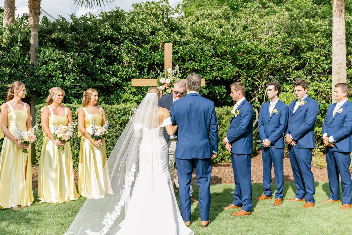 Outdoor wedding ceremony with bridal party wearing yellow dresses and navy suits standing under palm trees.