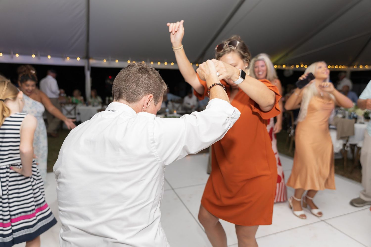People dancing with raised arms under a white tent at an outdoor celebration.