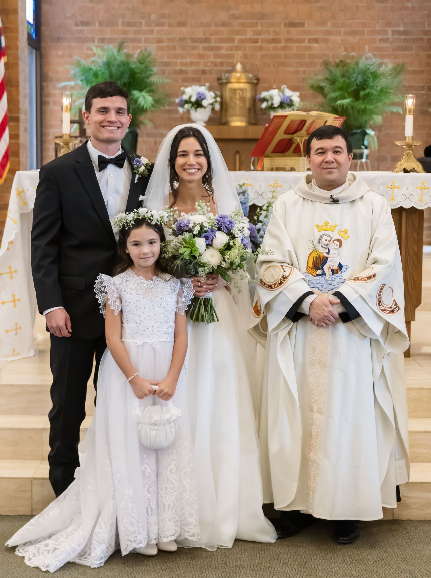 A group in formal attire including white dresses and tuxedos stands together in a church for a religious ceremony.