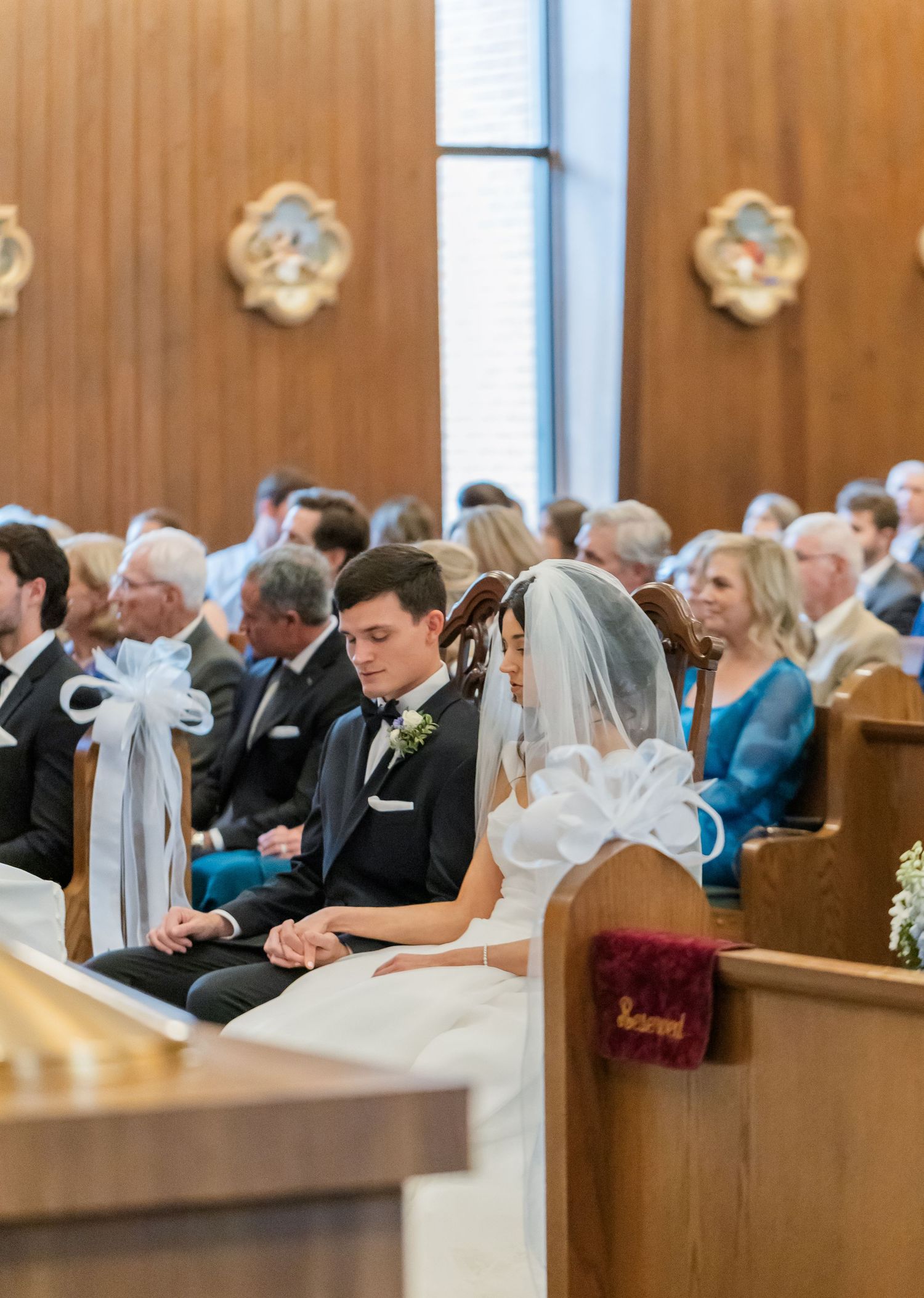 Wedding guests seated in wooden church pews decorated with white bows during ceremony.