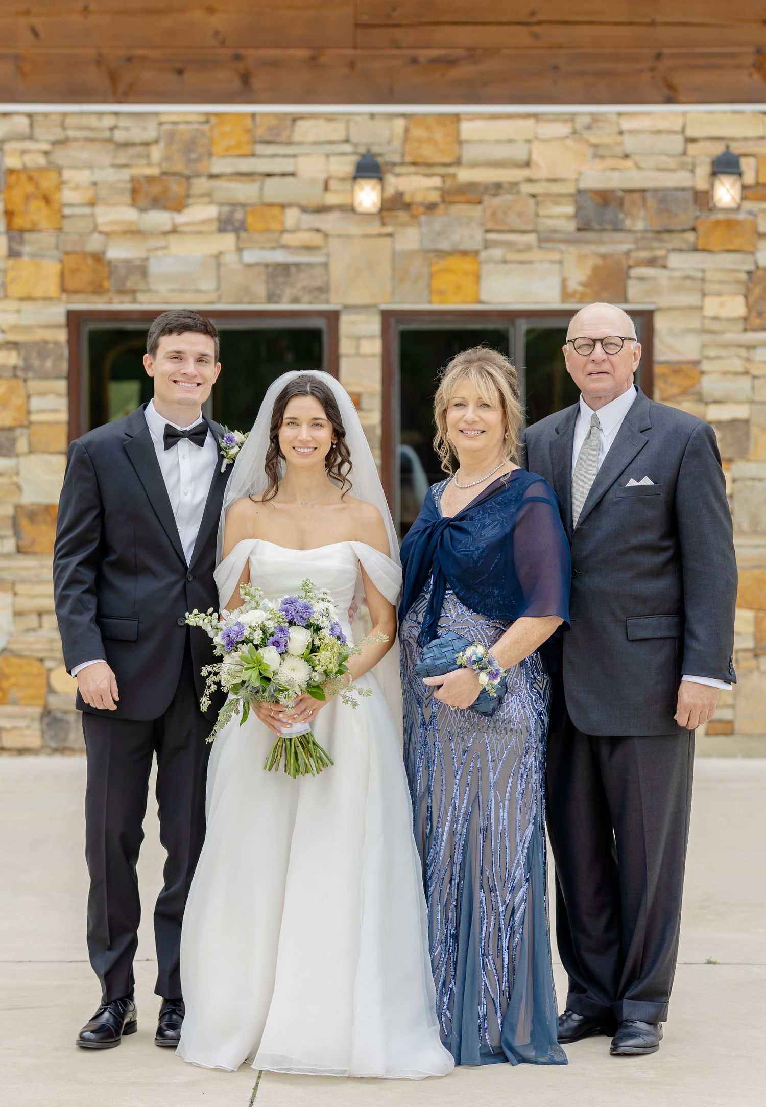 Wedding party in formal attire poses outside stone building, with bride holding purple and white floral bouquet.