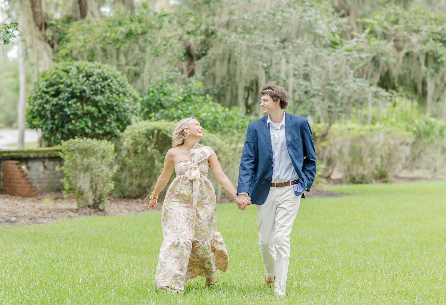 A couple walks hand in hand through a lush garden setting with Spanish moss hanging from trees in the background.