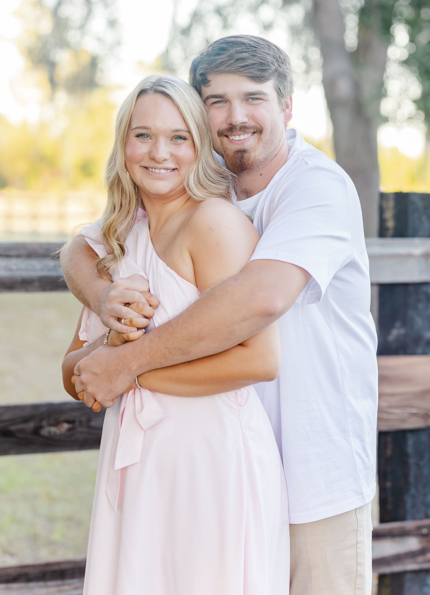 A couple in light-colored outfits embraces while posing outdoors in a natural setting with trees in the background.