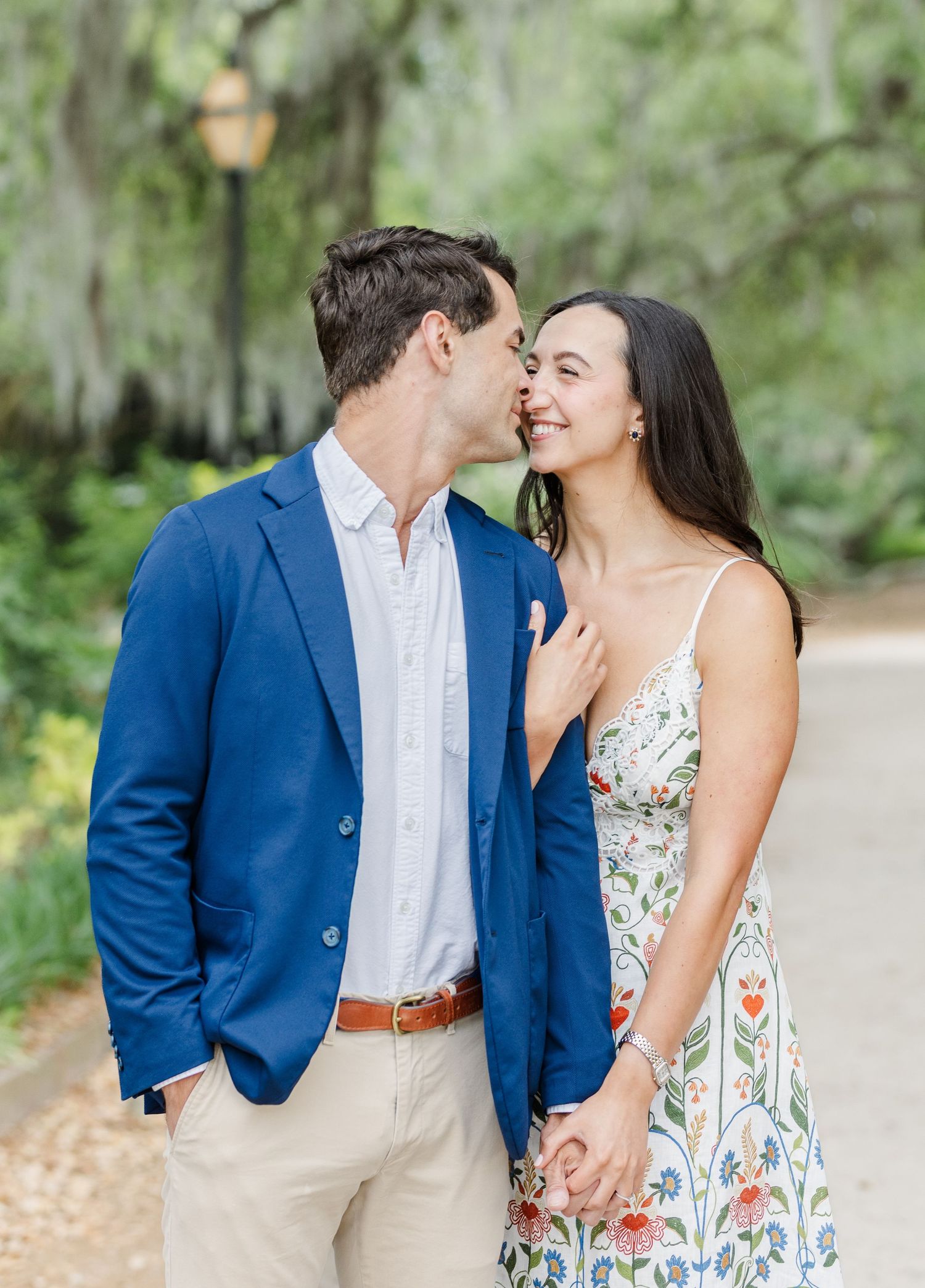 A couple shares a romantic moment outdoors, man in blue blazer kissing woman's cheek as she smiles, holding hands.