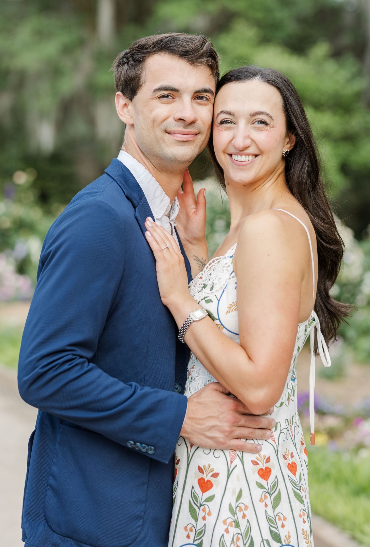 Smiling couple in close embrace, woman in floral dress and man in navy blazer, engagement portrait.