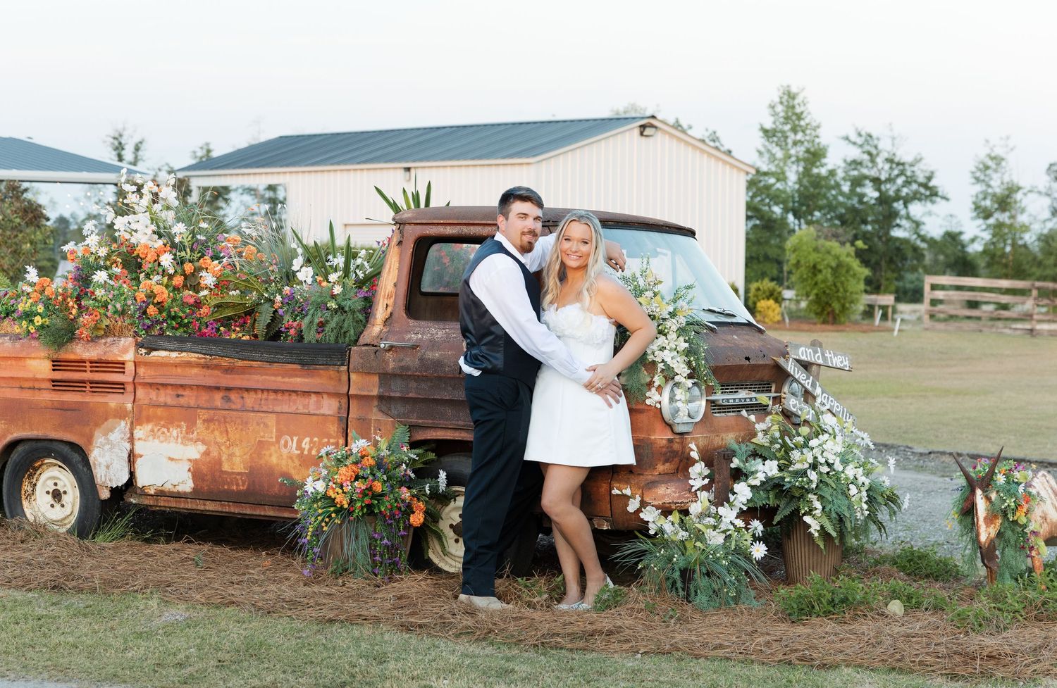Bride and groom posing beside a rustic truck decorated with colorful floral arrangements.