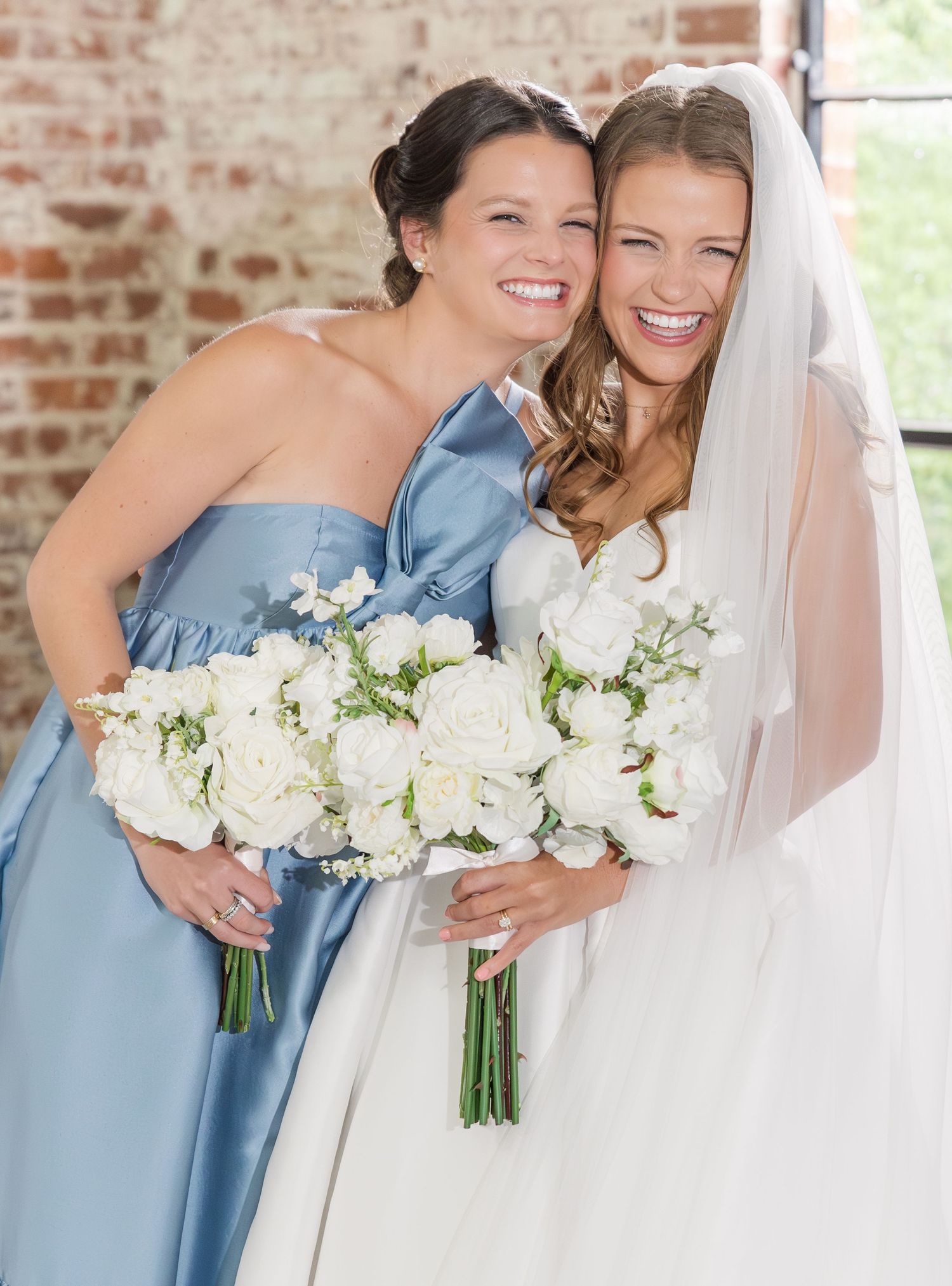 Smiling bride in white gown and veil with bridesmaid in blue dress holding white rose bouquets.