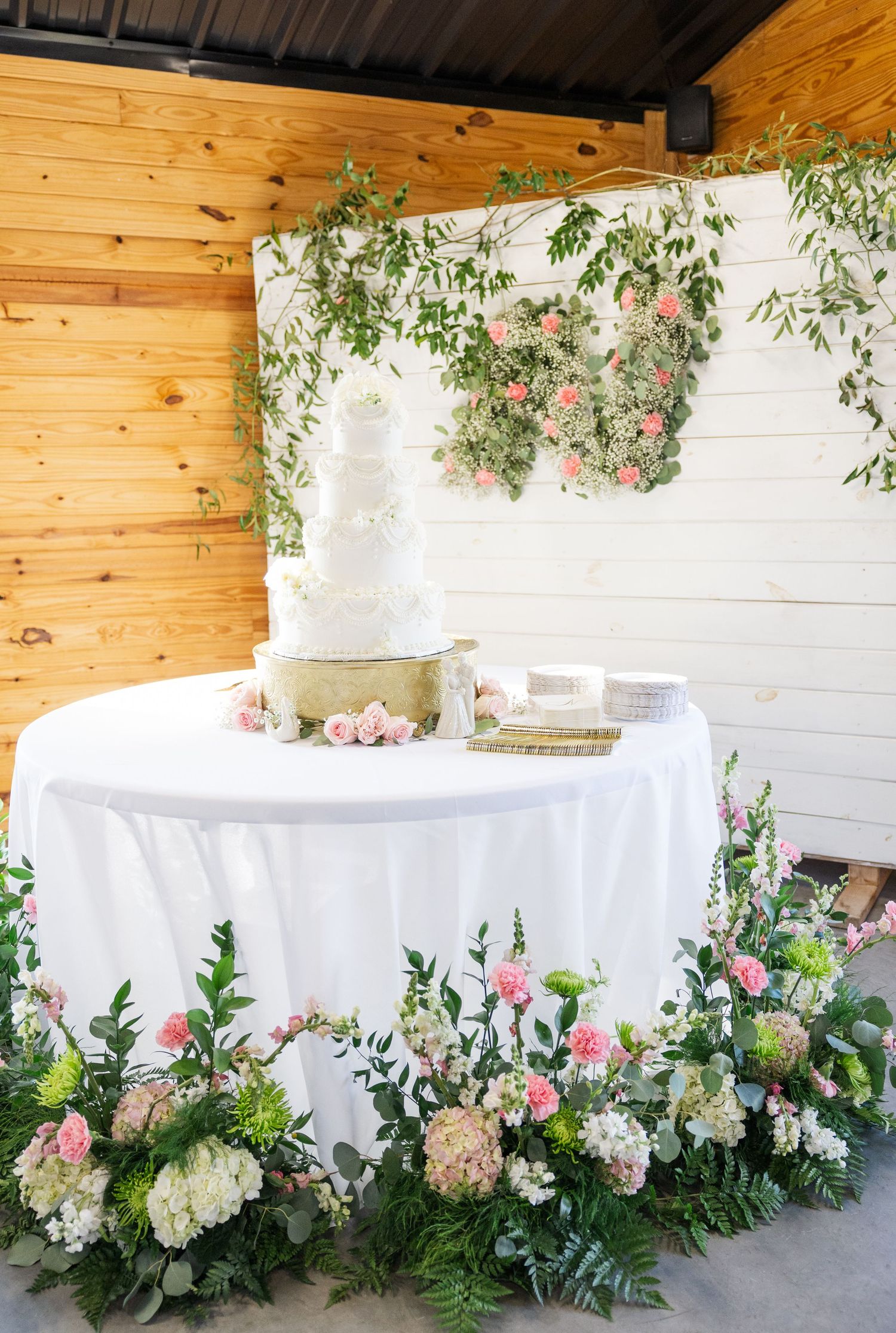 Four-tier white wedding cake on draped table with lush pink and white floral arrangements and greenery backdrop.