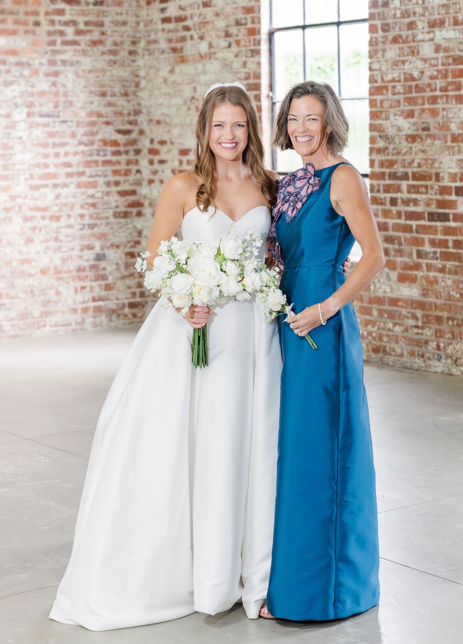 Bride in white gown holding white bouquet poses with woman in blue dress against brick wall.