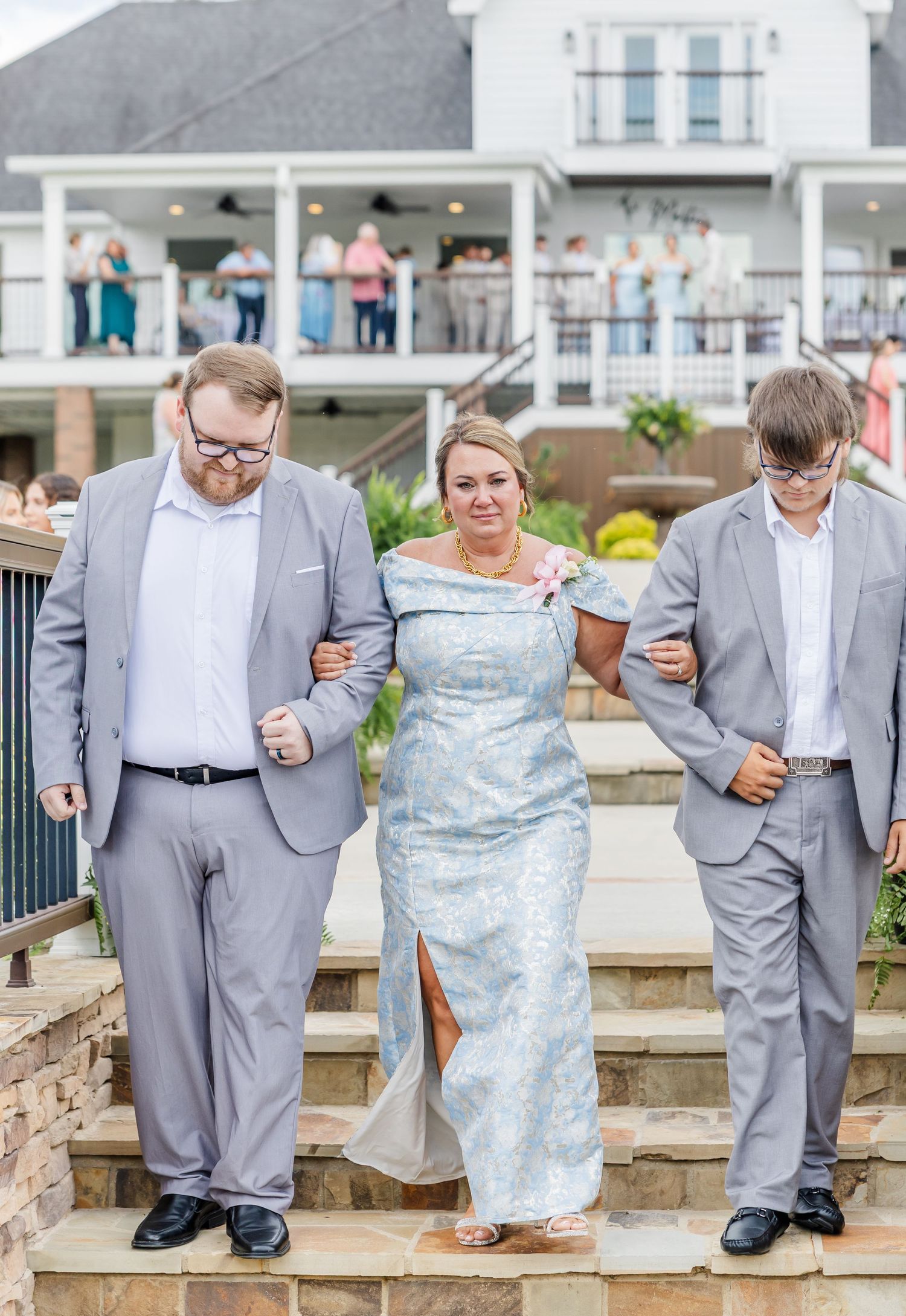 A woman in a blue gown walks down stone steps escorted by two men in gray suits at an outdoor wedding venue.