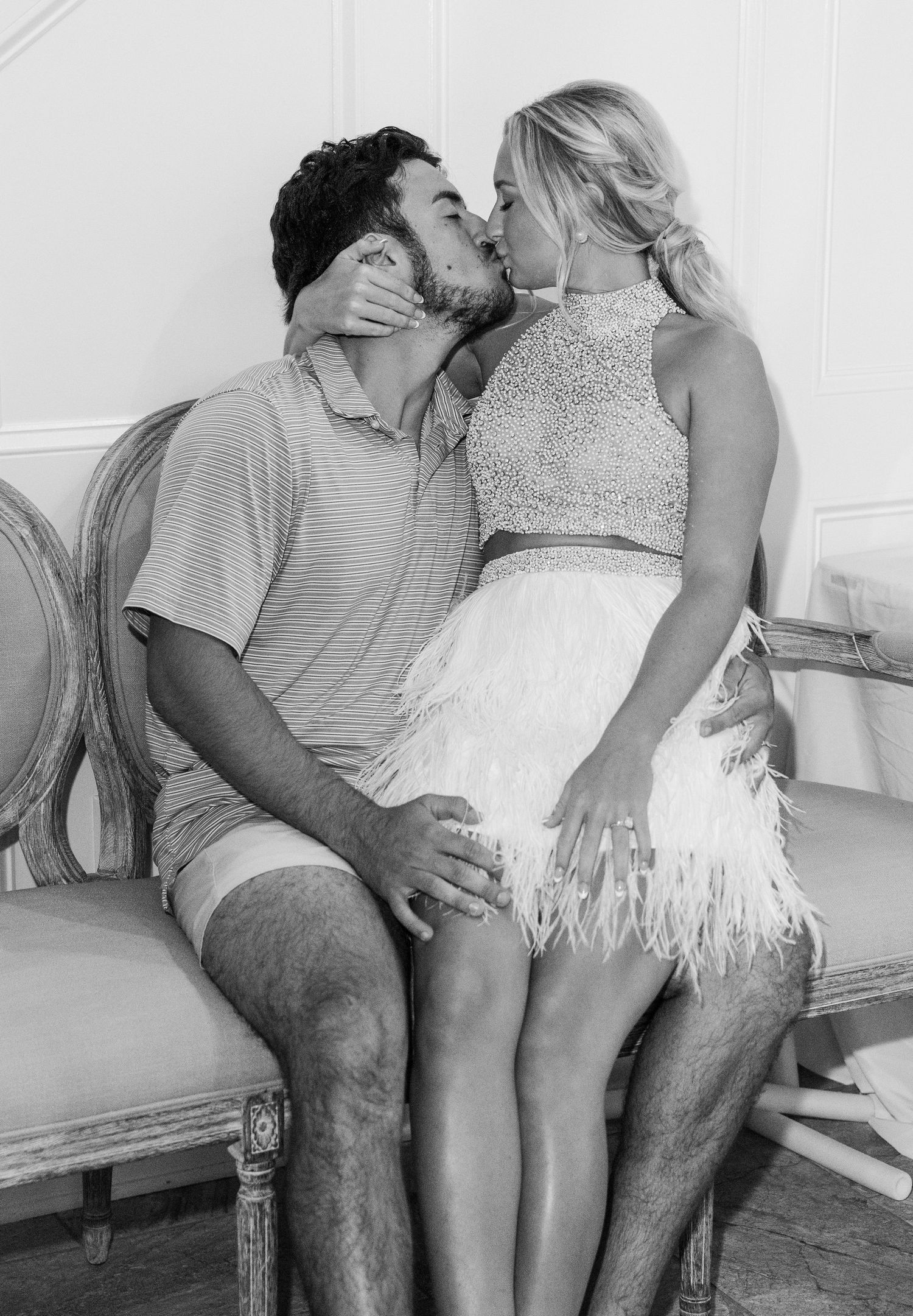 Couple shares romantic kiss while seated on ornate French chair in black and white photo.