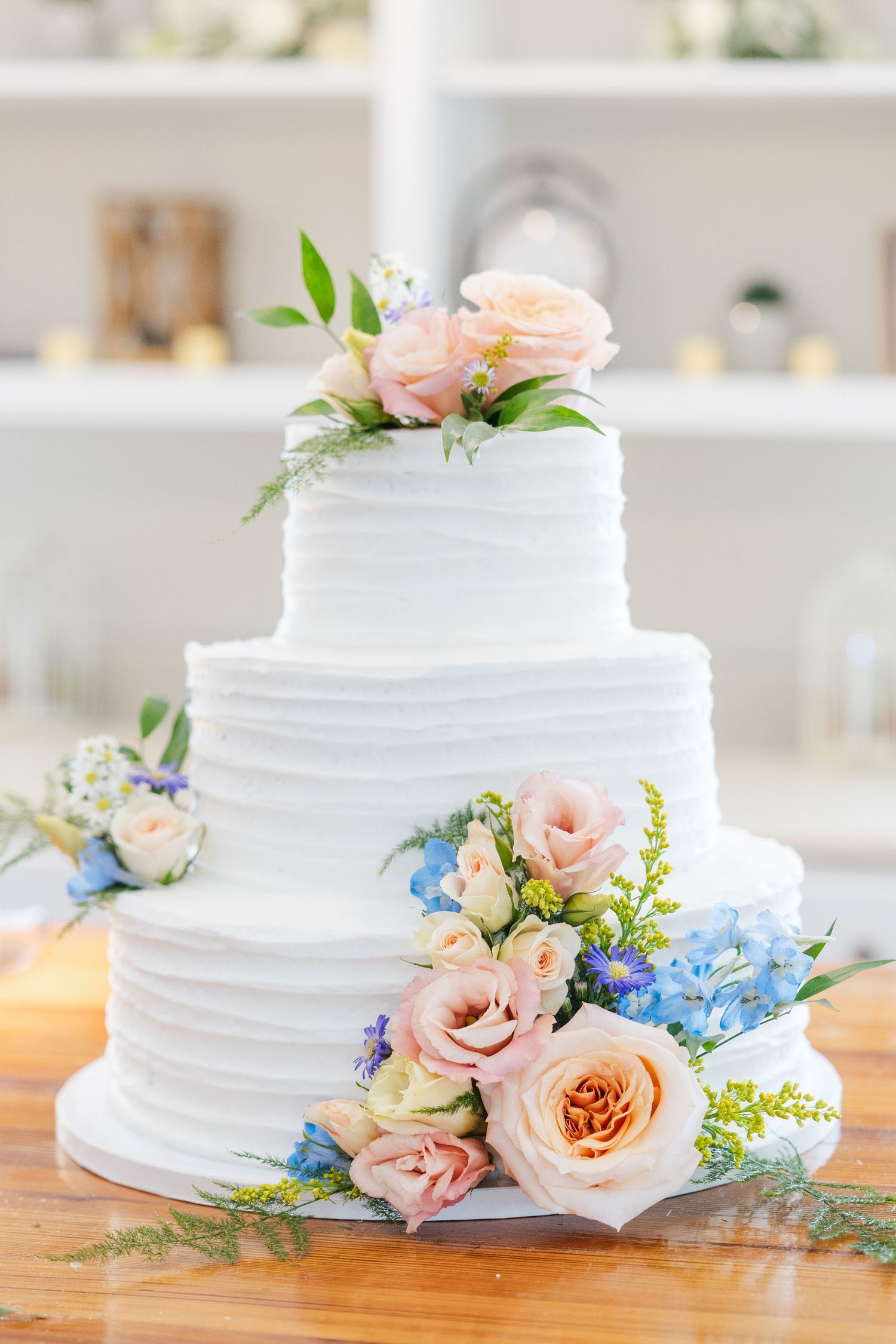 Three-tier white wedding cake with textured frosting adorned with pink roses and blue hydrangeas.