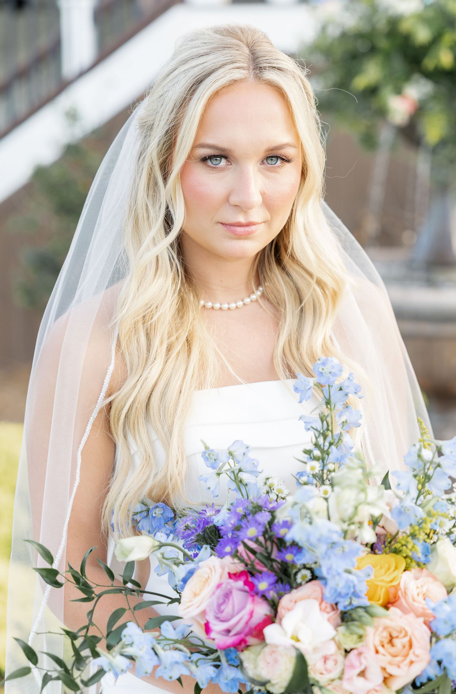 Blonde bride wearing a veil and pearl necklace holds a vibrant blue and pink floral bouquet.