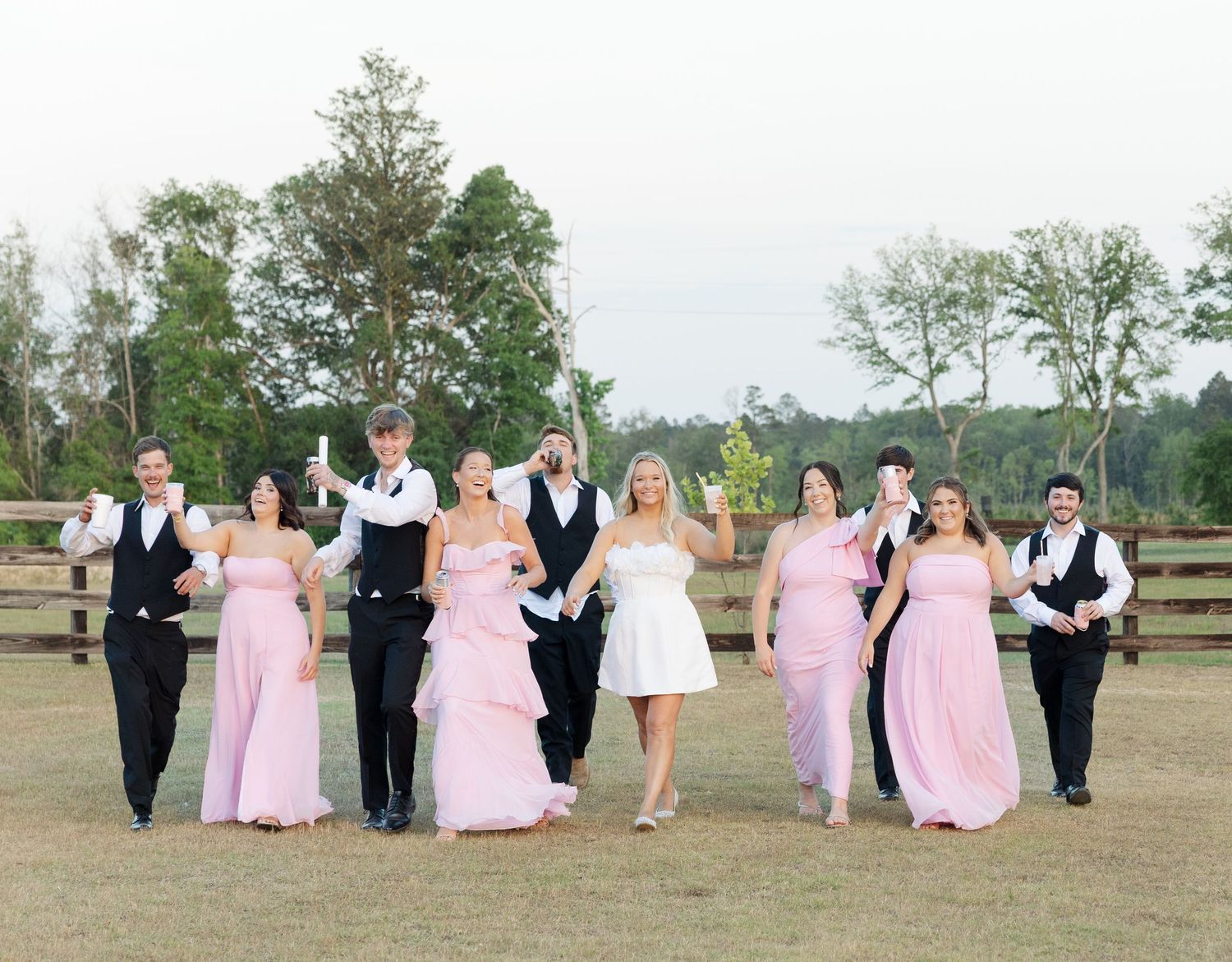 Wedding party in pink bridesmaid dresses and black tuxedos celebrating outdoors on a green lawn with wooden fence.