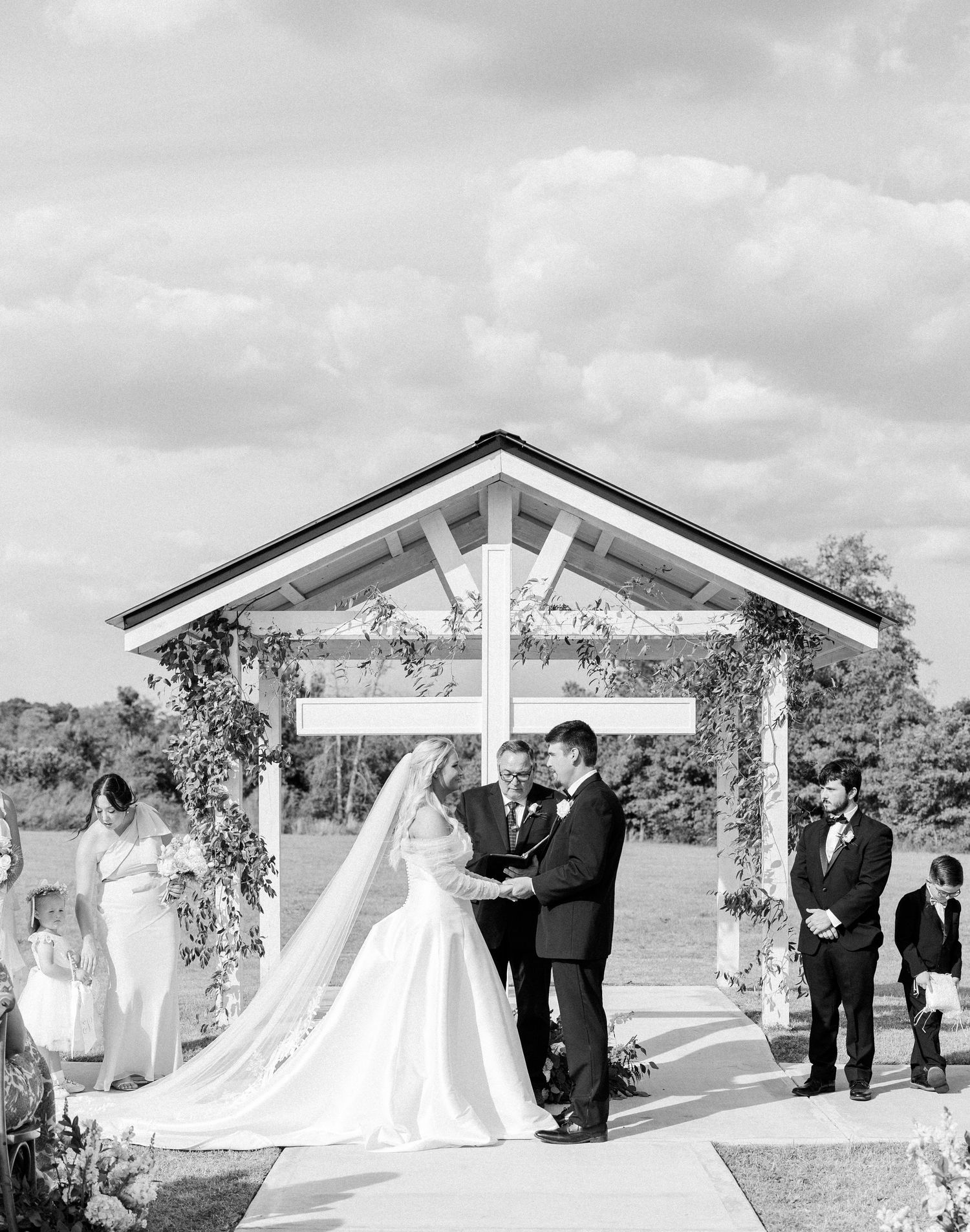Black and white photo of bride and groom exchanging vows under floral pergola outdoors.