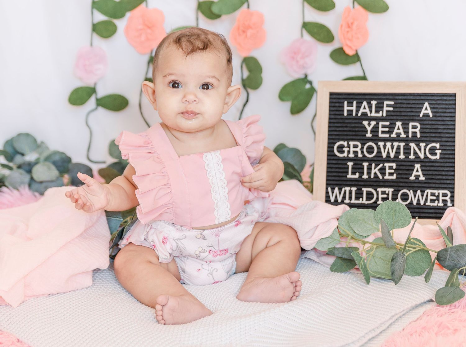 Baby in pink dress sitting beside blackboard sign against floral background.