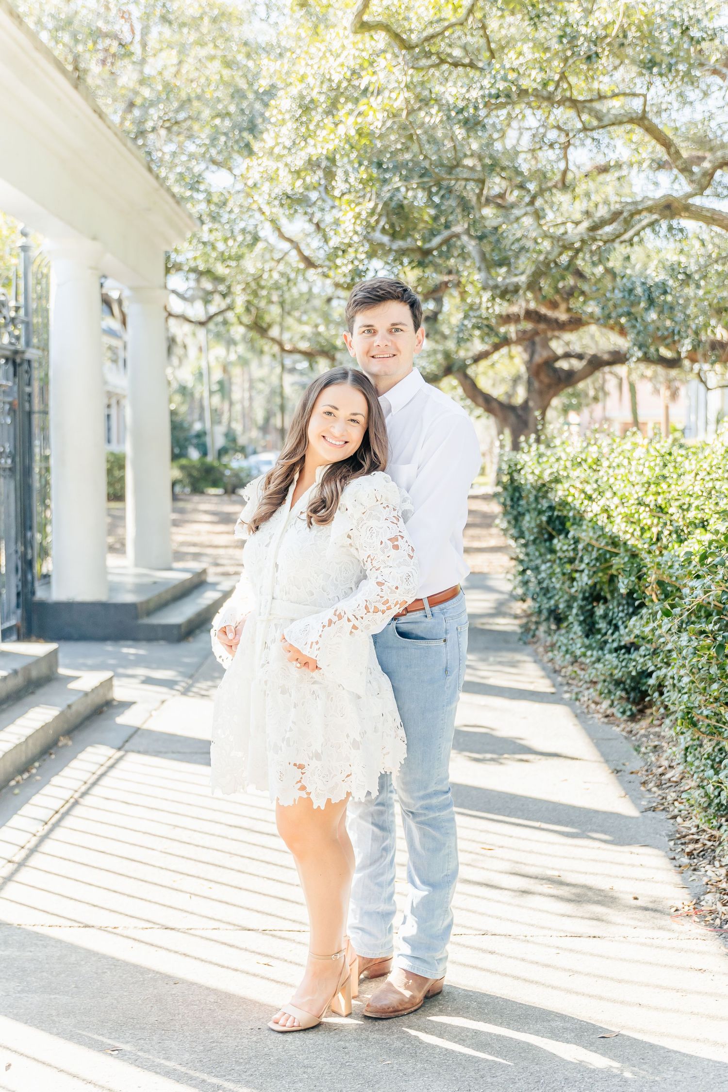 Sun-drenched engagement photo of couple in white and denim against garden columns.