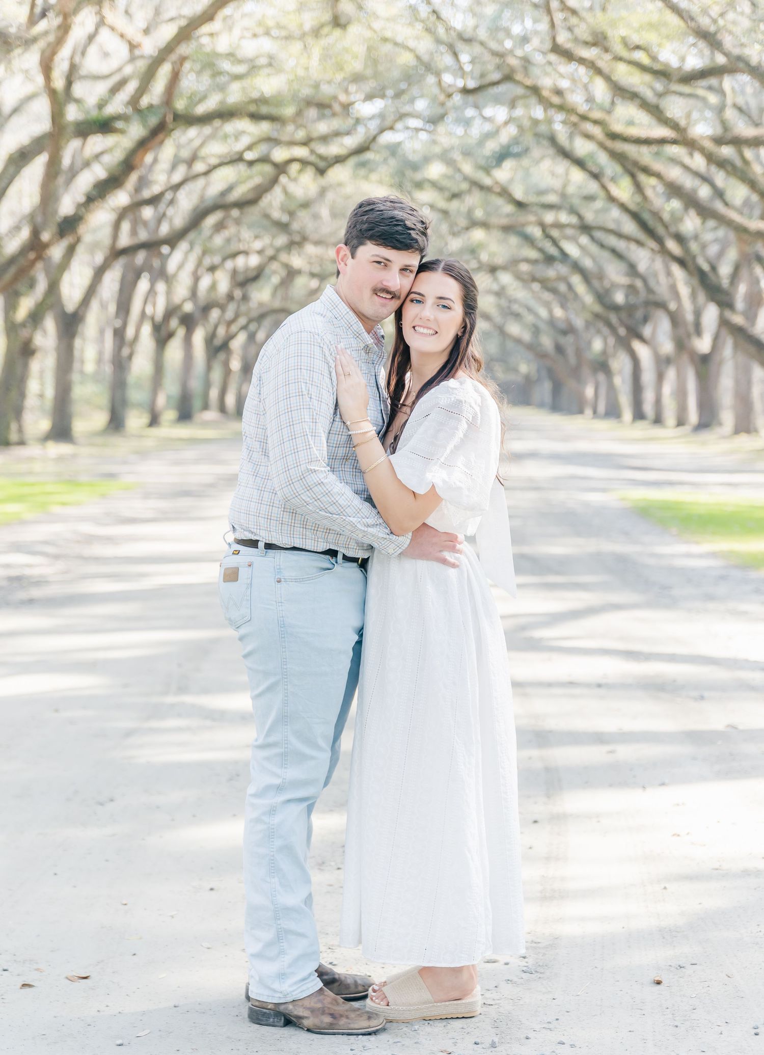 A couple in coordinated outfits poses lovingly on a scenic oak-lined pathway.