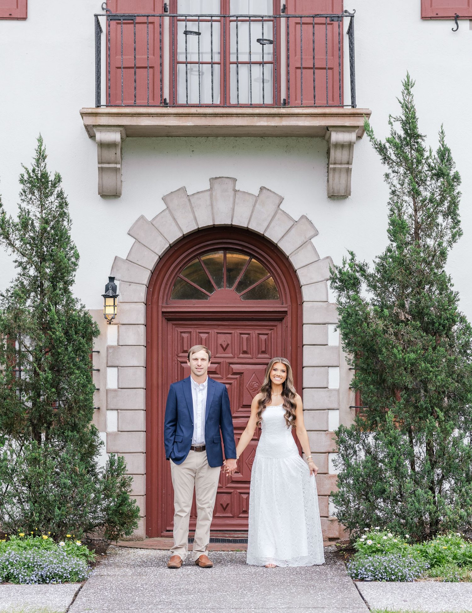 Couple holding hands in front of elegant red arched door with balcony, flanked by evergreen trees.
