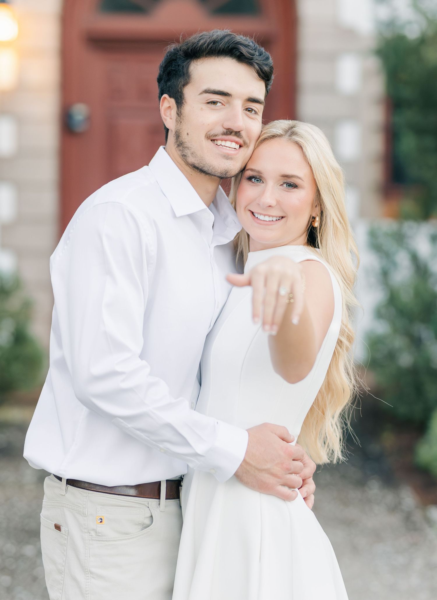 Smiling engaged couple in white outfits embracing, woman showing off diamond engagement ring to camera.