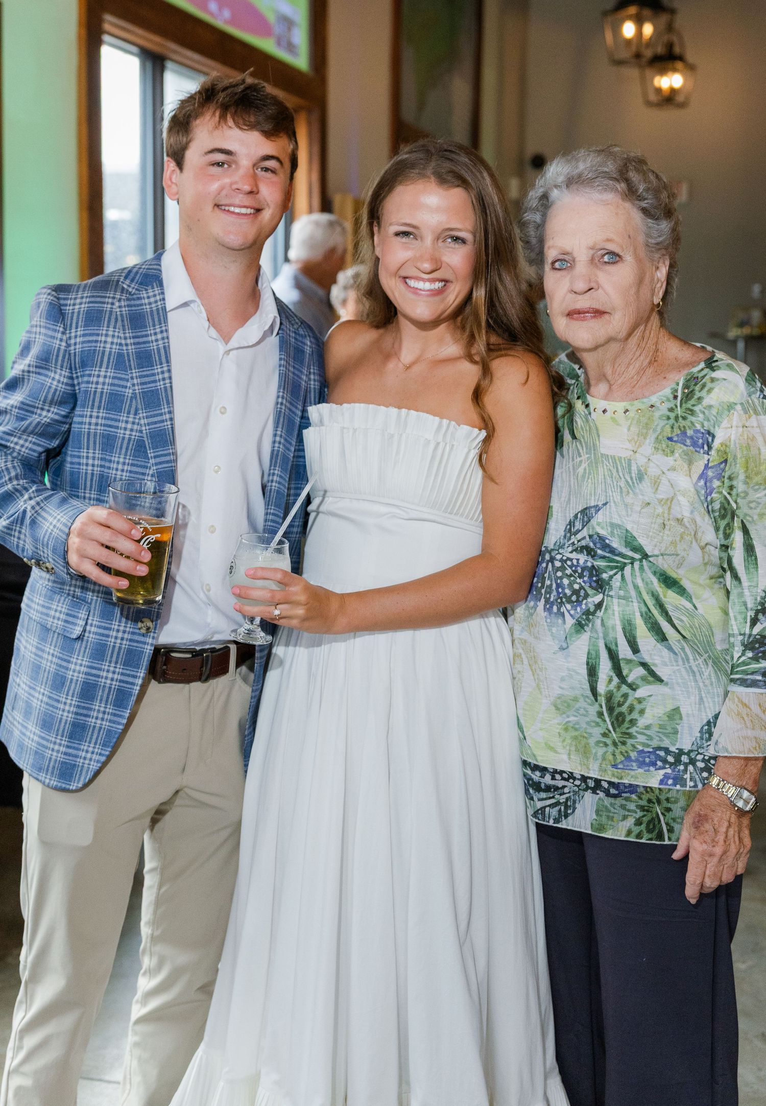 Young man in plaid blazer and bride in white dress pose with elderly woman at wedding reception.