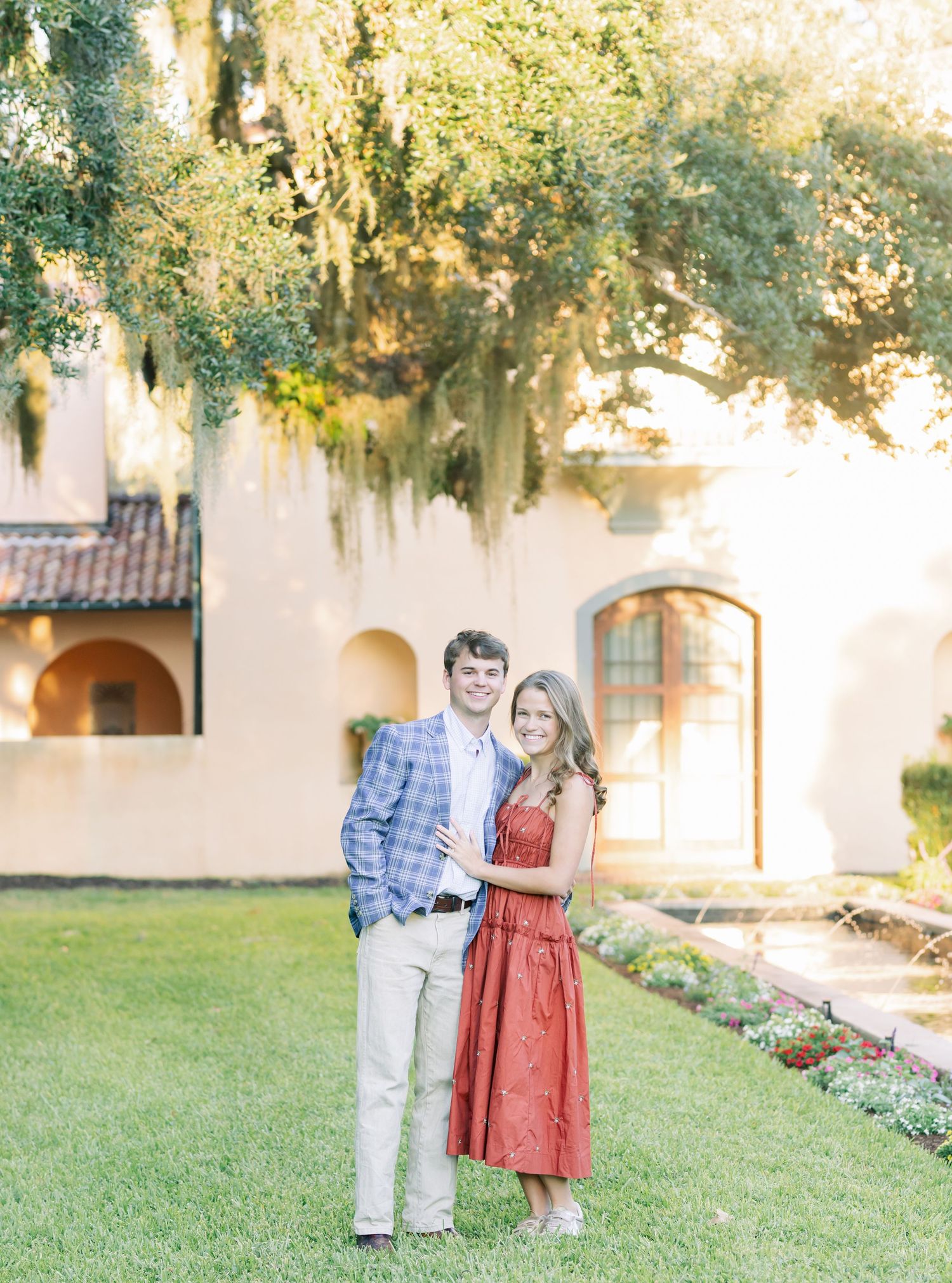 A couple walks hand in hand on a grassy path with Spanish architecture in the background.