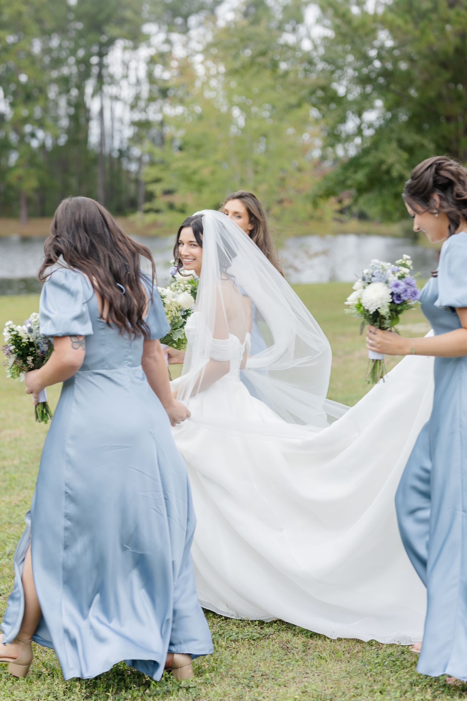 Bridesmaids in blue dresses help with bride's flowing veil on grassy lawn.