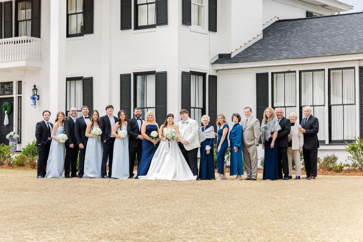 Wedding party and family group photo in front of elegant white venue with black shutters on sunny wedding day.