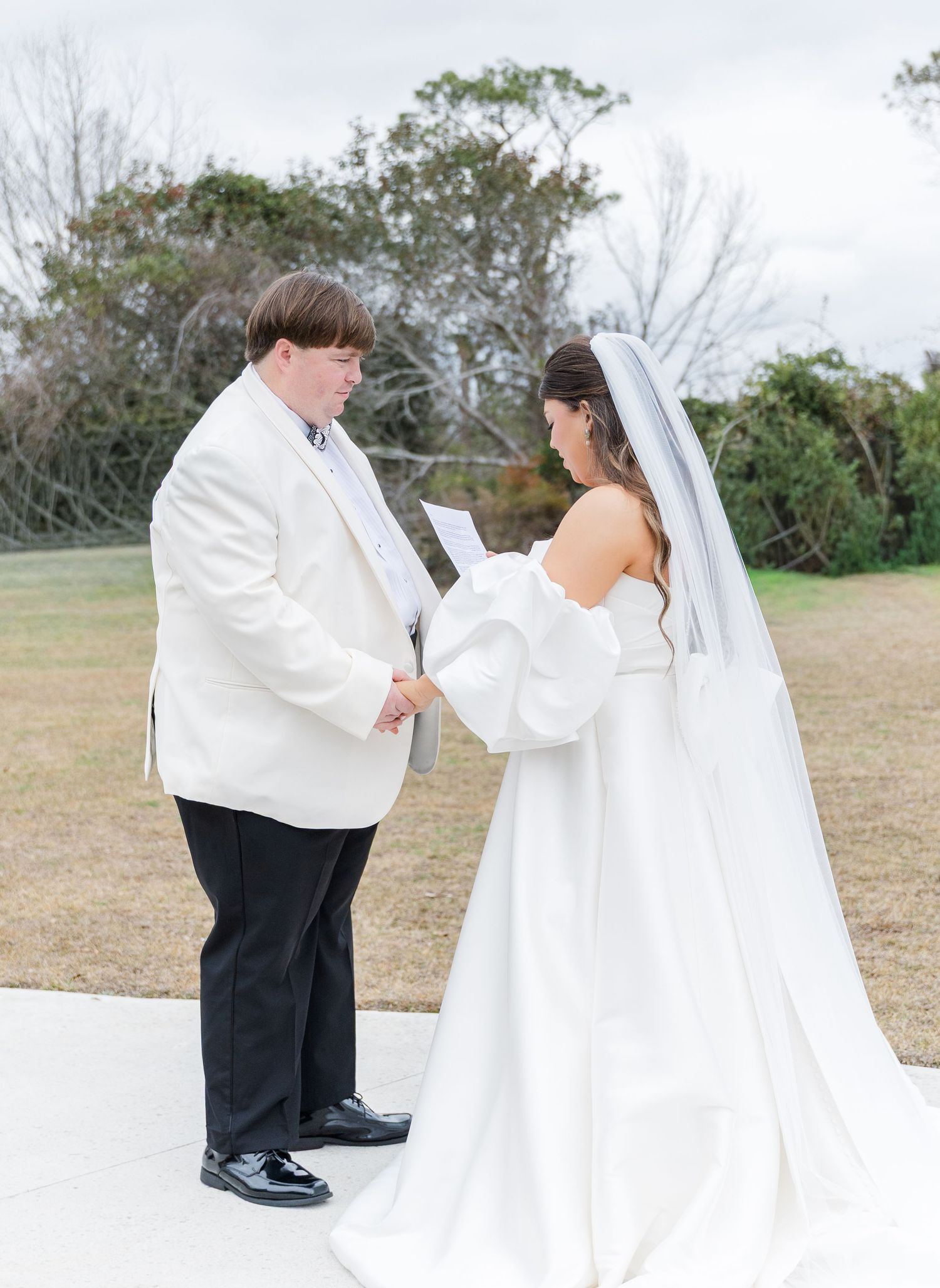 Groom in white jacket reads vows to bride in off-shoulder gown with cathedral veil outdoors.