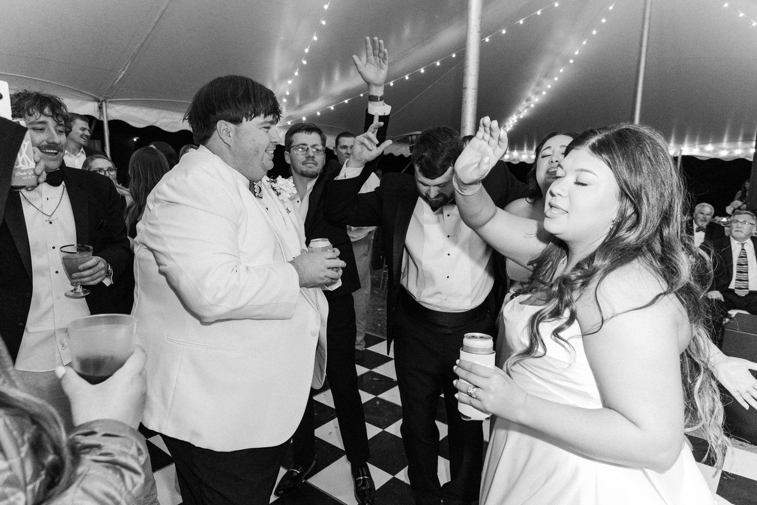 Guests dancing and celebrating in black and white checkered dance floor under tent at wedding reception.
