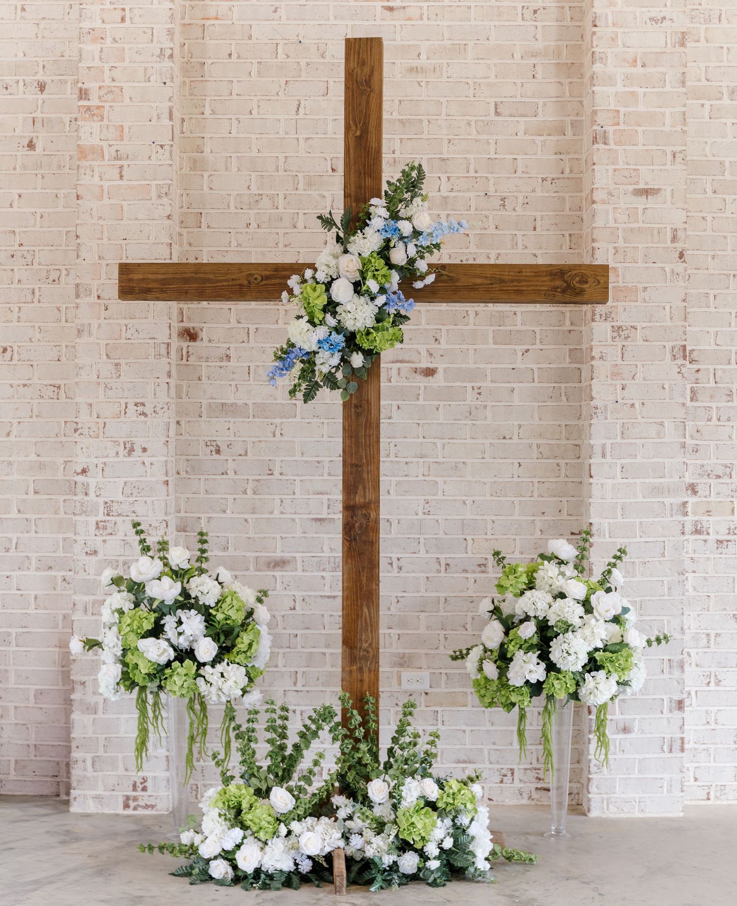 Rustic wooden cross adorned with white and blue floral arrangements against a white brick wall for a wedding ceremony.