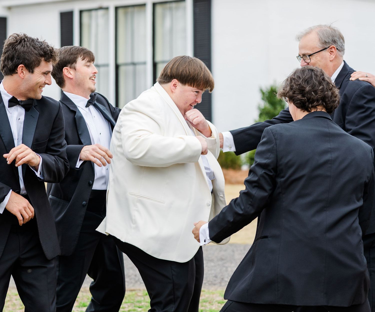 Groom in white tuxedo jacket laughing with groomsmen in black tuxedos outdoors.