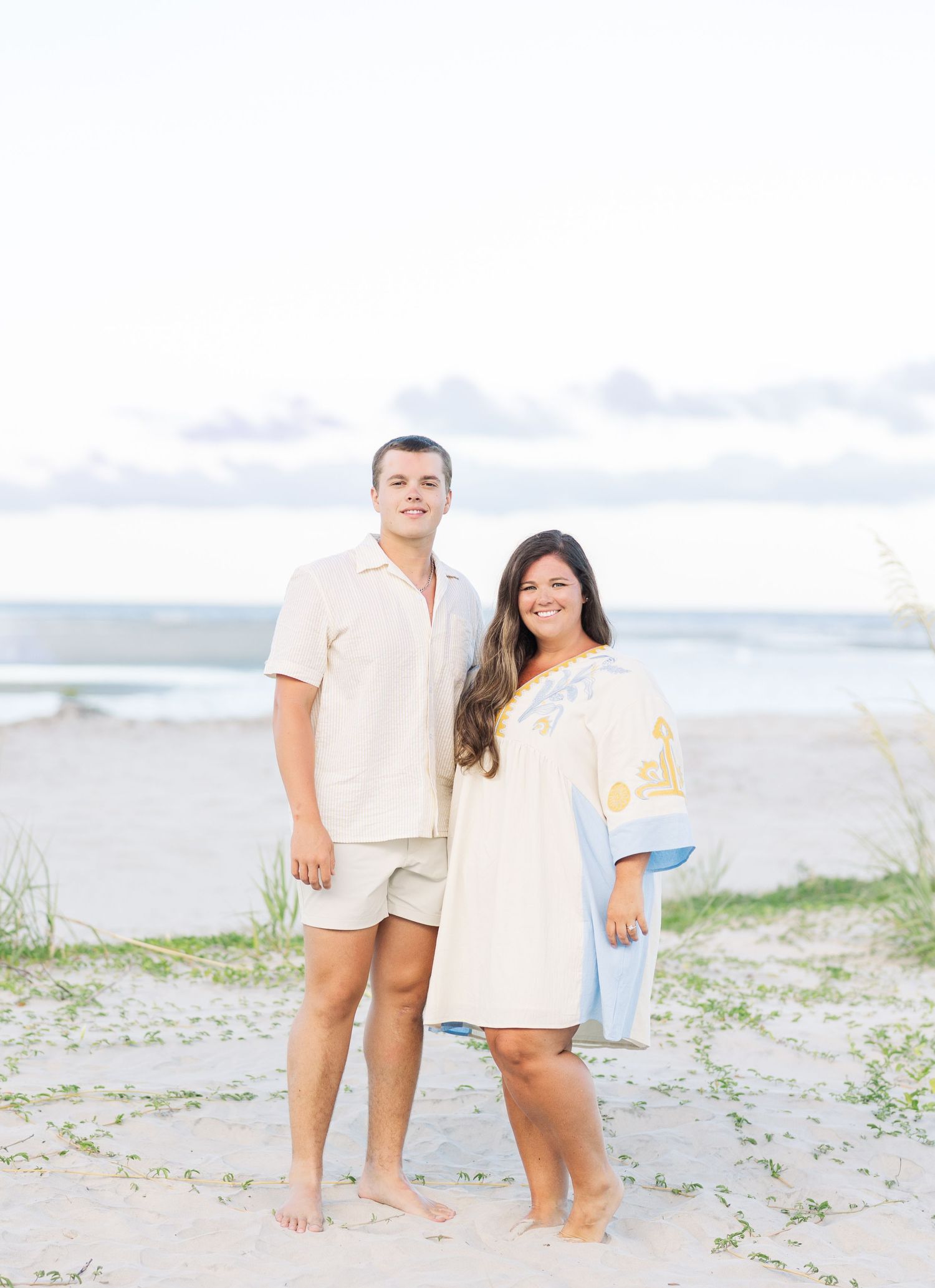 A couple stands barefoot on a sandy beach wearing light colored outfits against an ocean backdrop.
