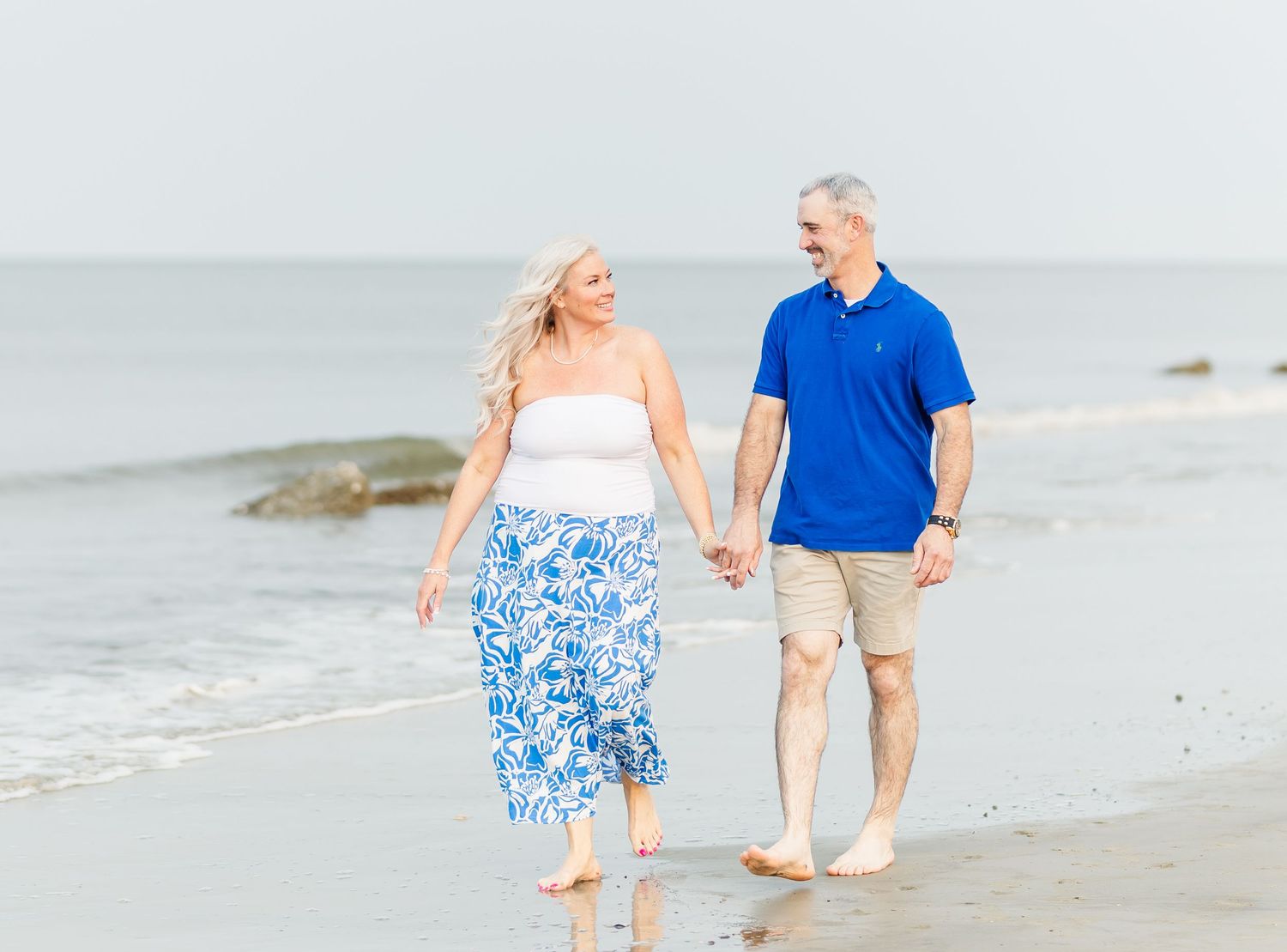 A couple walking hand in hand along a sandy beach wearing coordinated blue and white outfits.