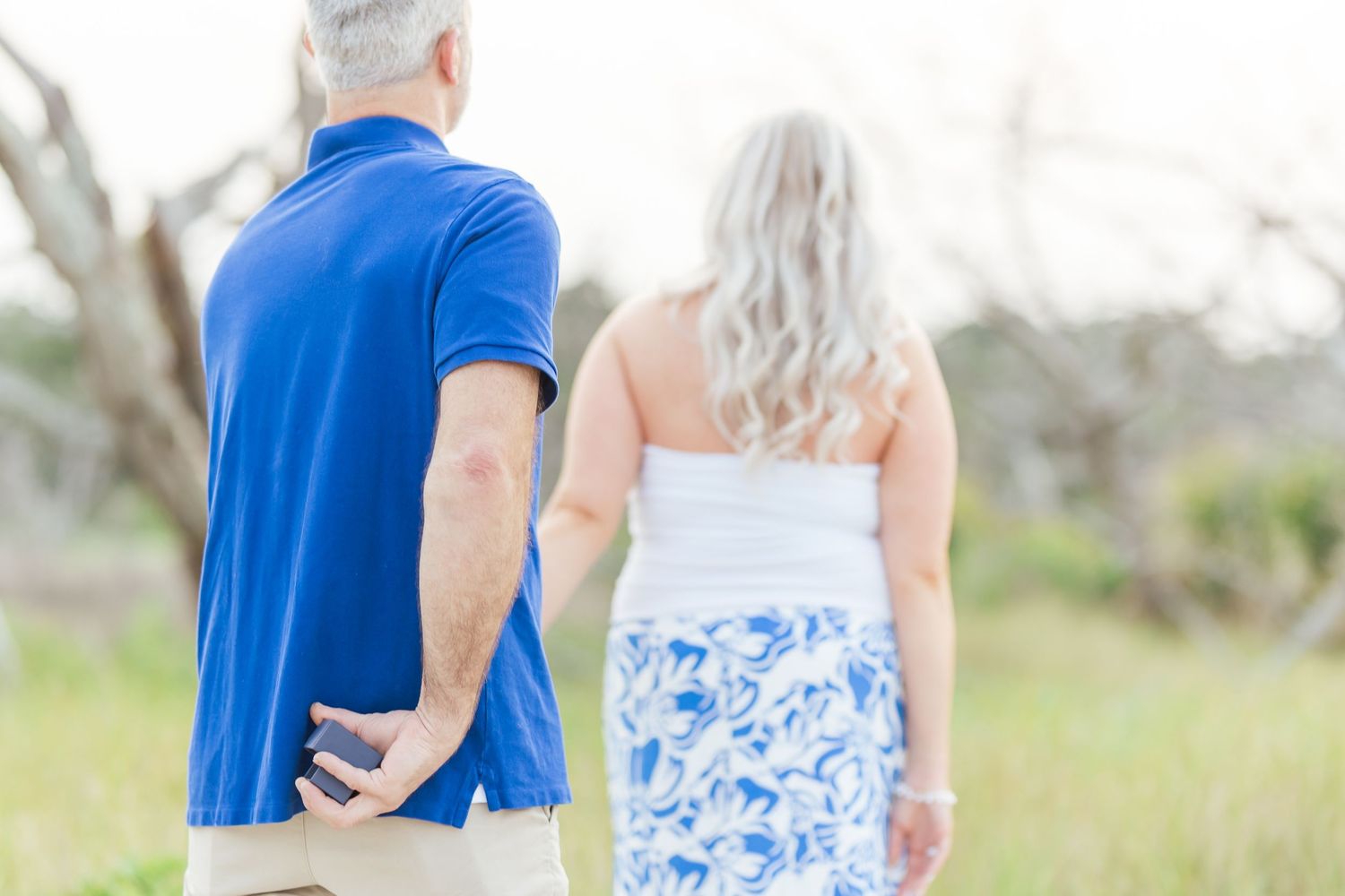 A couple wearing blue and white clothing holds hands while standing in a grassy meadow on a bright sunny day.