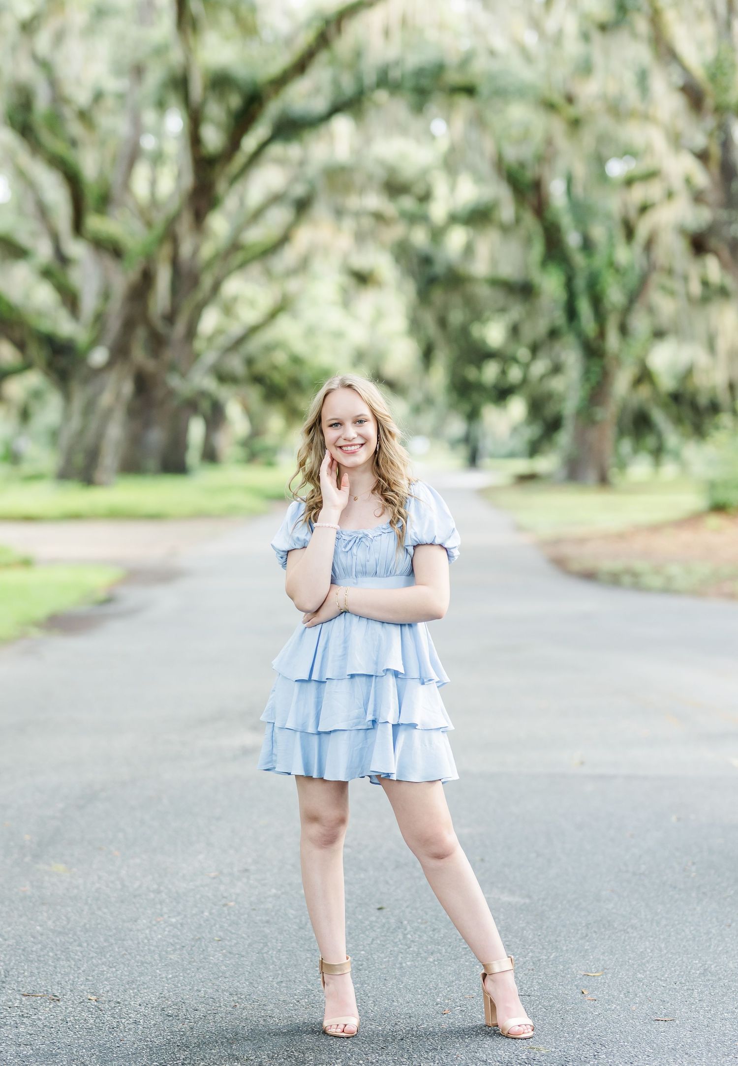 A person in a light blue tiered ruffle dress poses confidently on a tree-lined pathway outdoors.