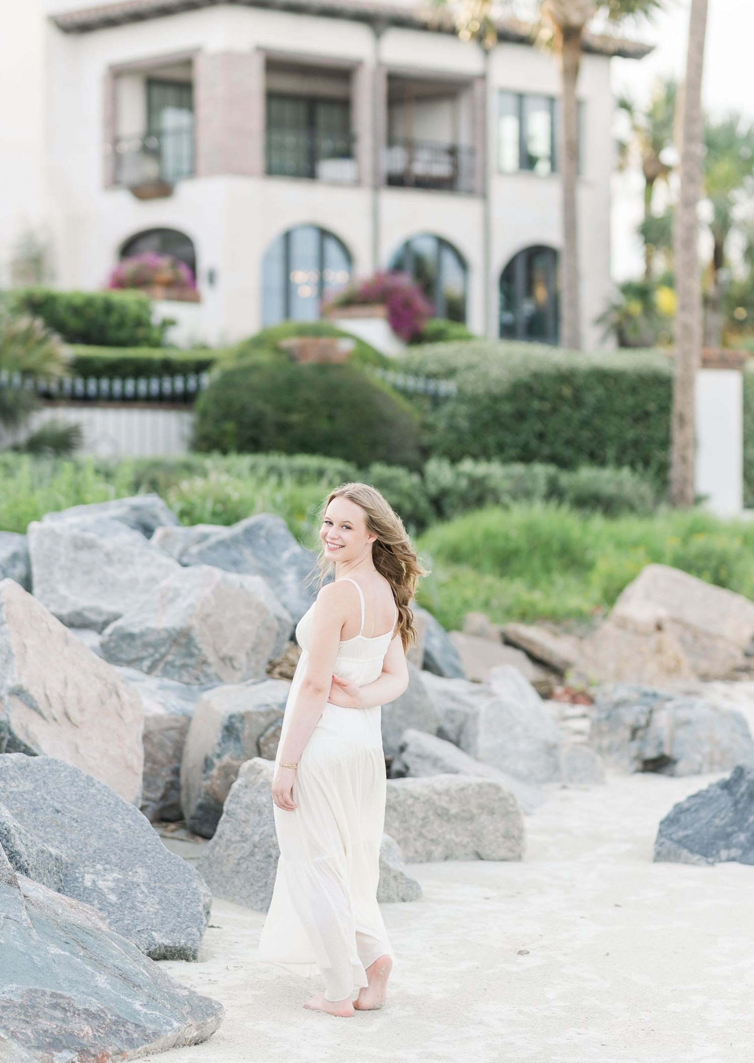 A flowing white dress contrasts against rocky coastal landscape with Mediterranean architecture in background.