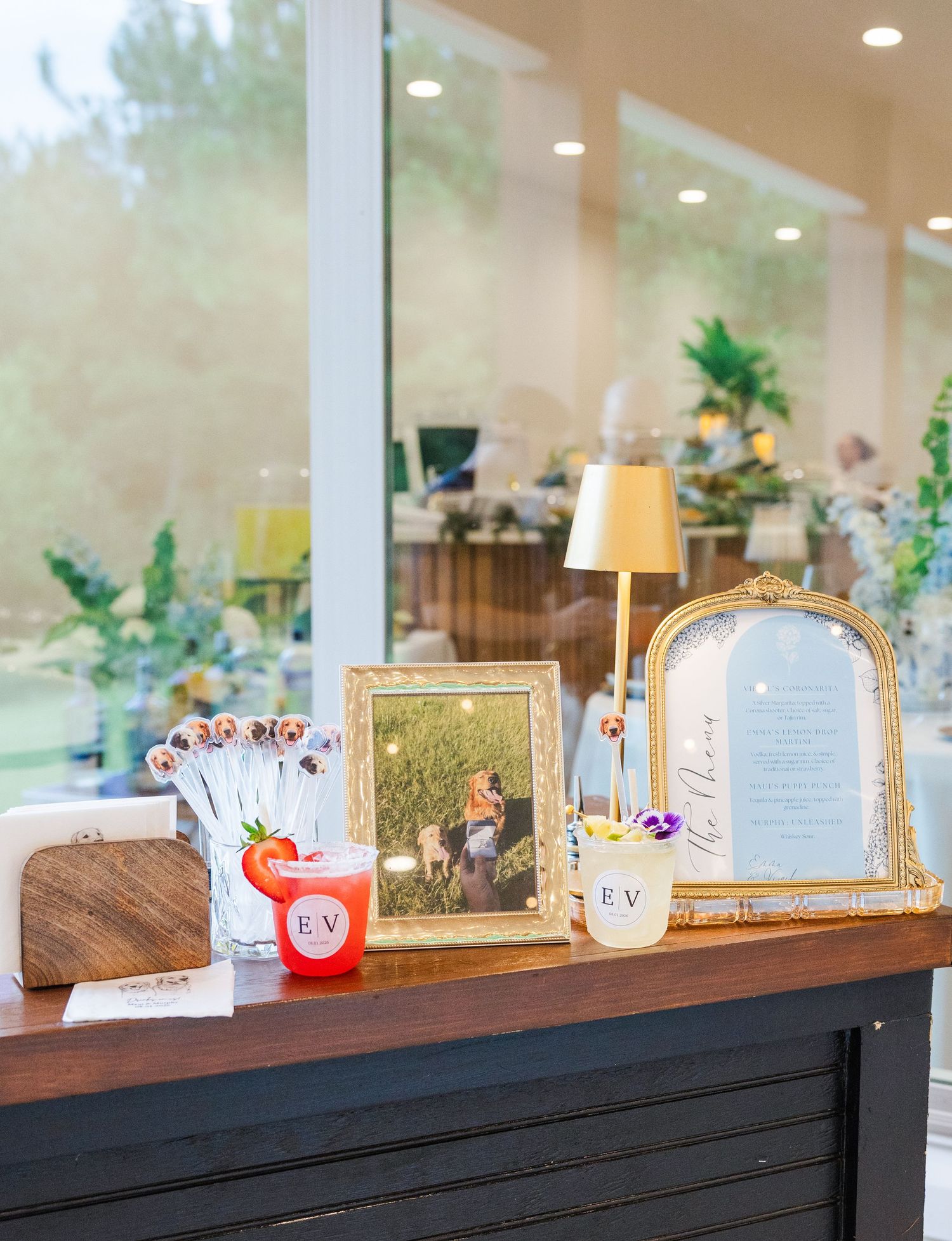 Elegant wedding bar display with monogrammed cocktails, gold frames, couple photo, and decorative signage at an upscale venue.