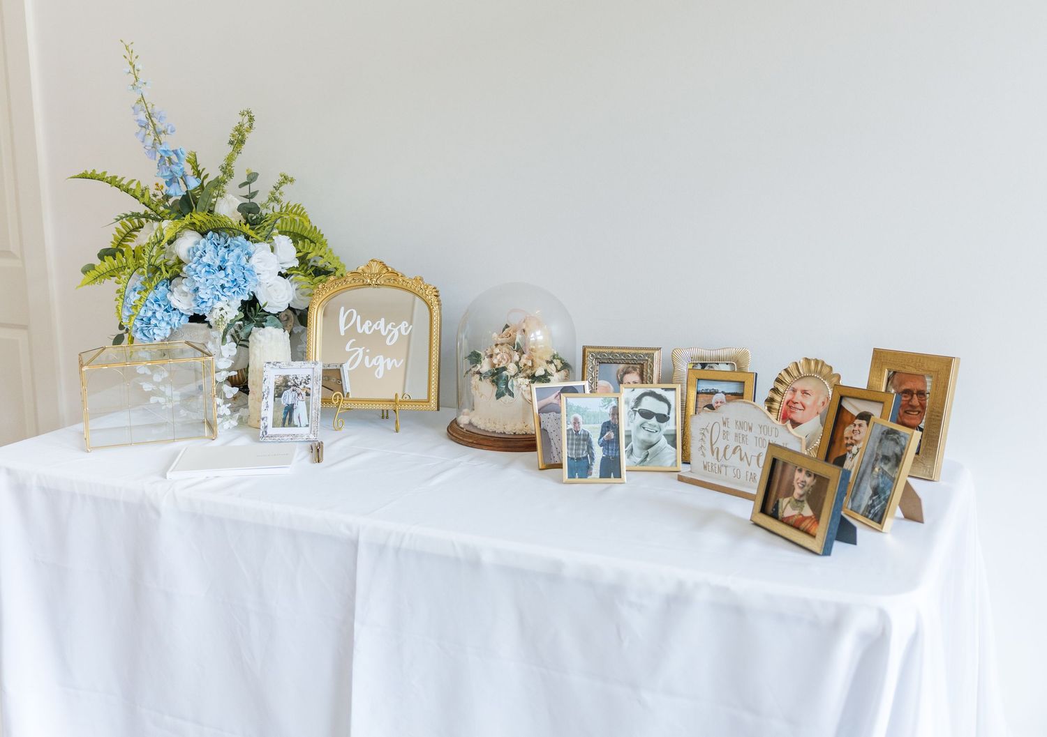 Elegant wedding memory table with framed photos, blue hydrangeas, and gold accents on a white linen tablecloth.