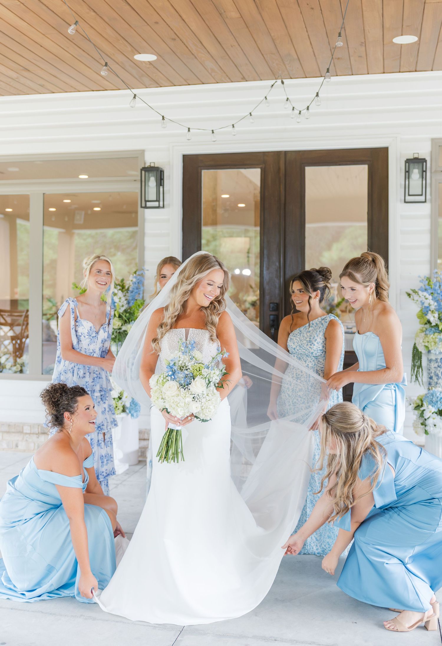 Bride in white gown and veil surrounded by bridesmaids in blue dresses on a covered porch during wedding day preparations.