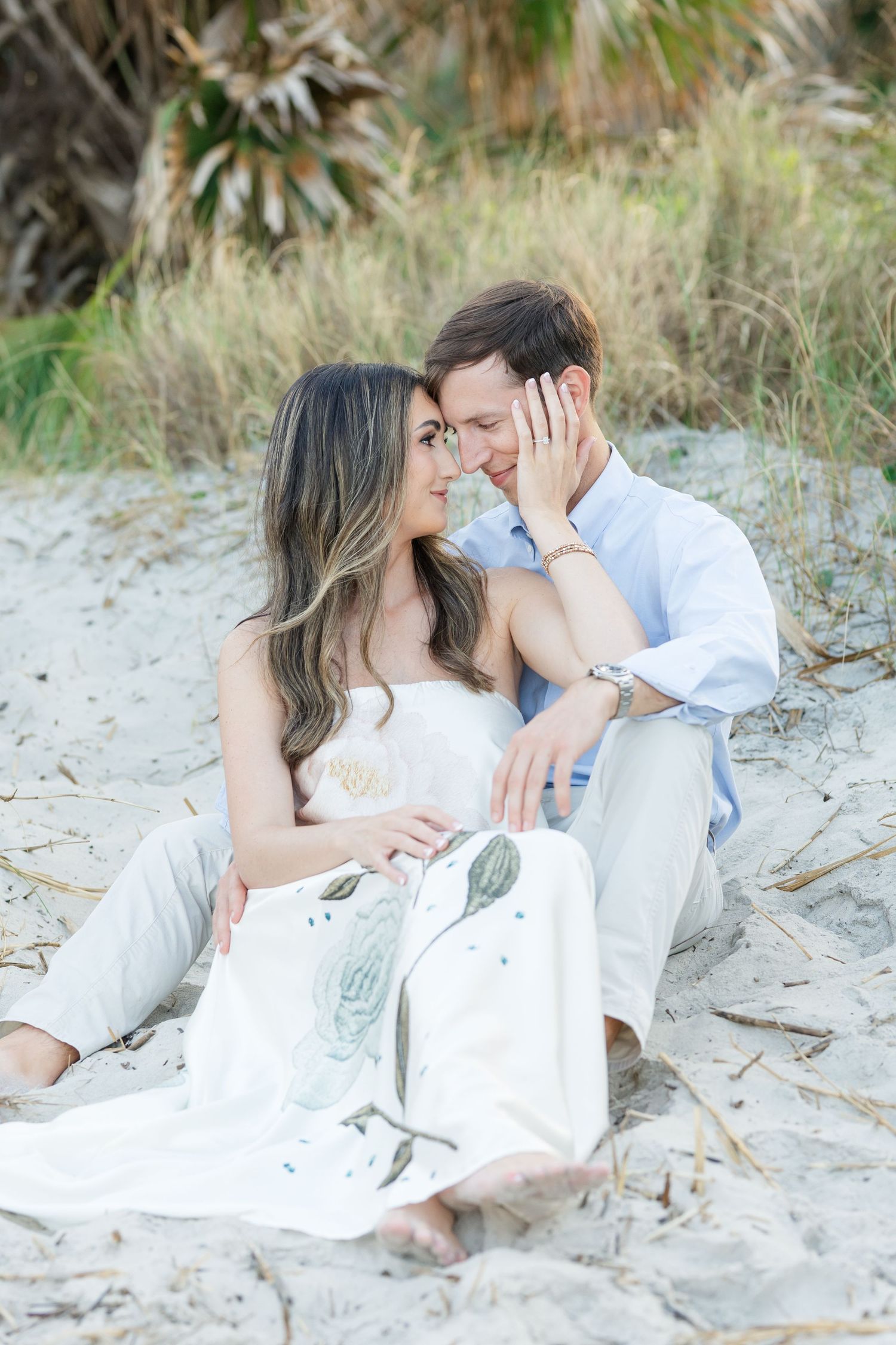 Romantic couple sitting on beach sand, smiling at each other, woman in white floral dress, man in blue shirt.