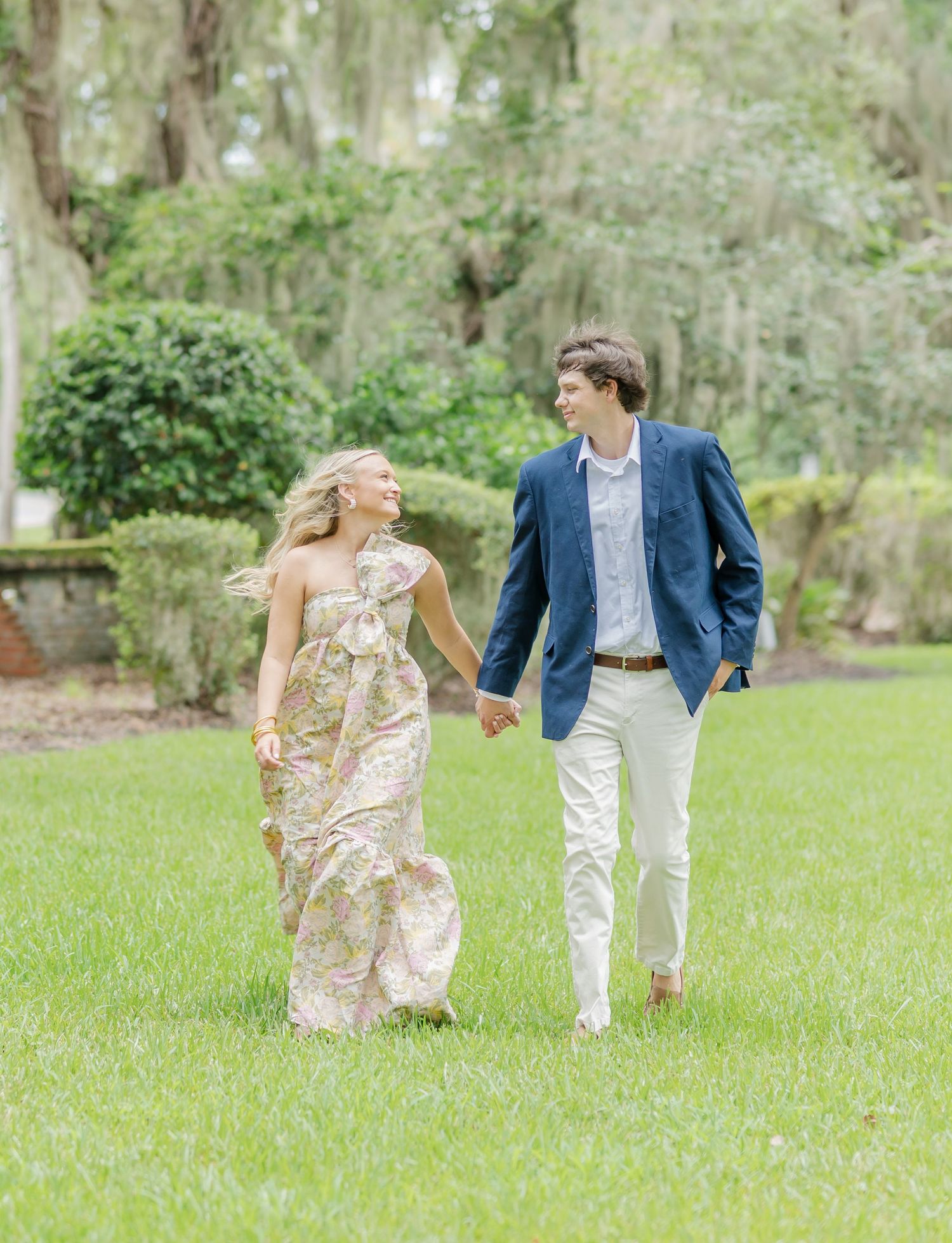 A happy couple strolls on green grass with Spanish moss-draped trees in the background.