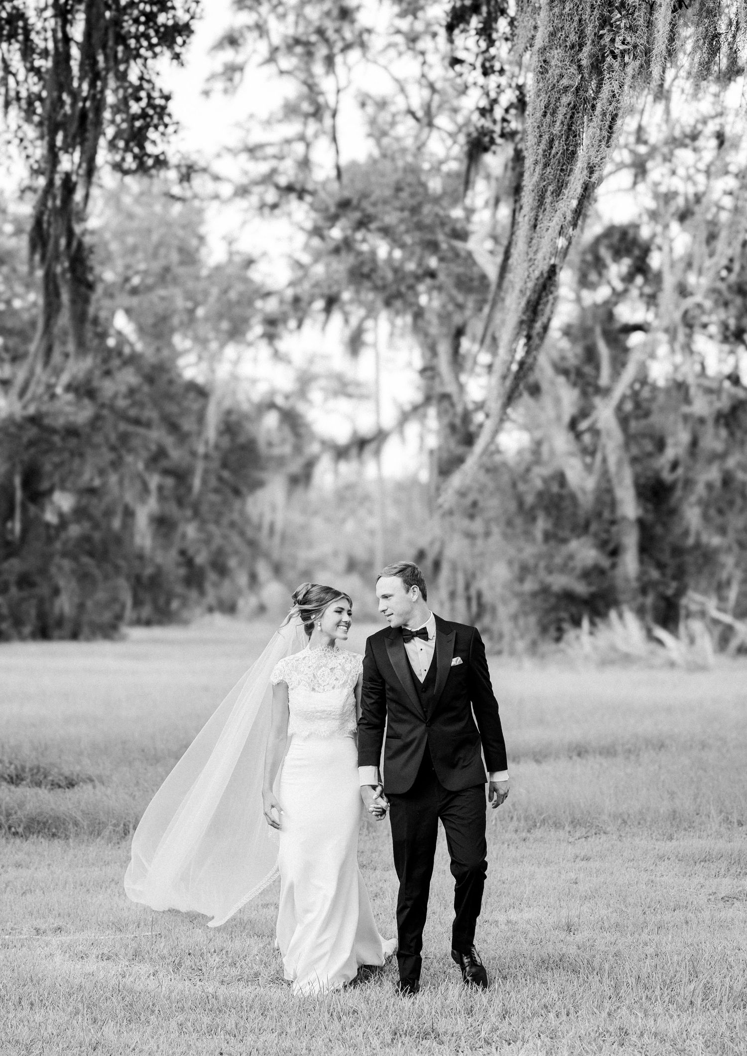 Black and white photo of bride and groom holding hands in a grassy field under Spanish moss-draped oak trees.