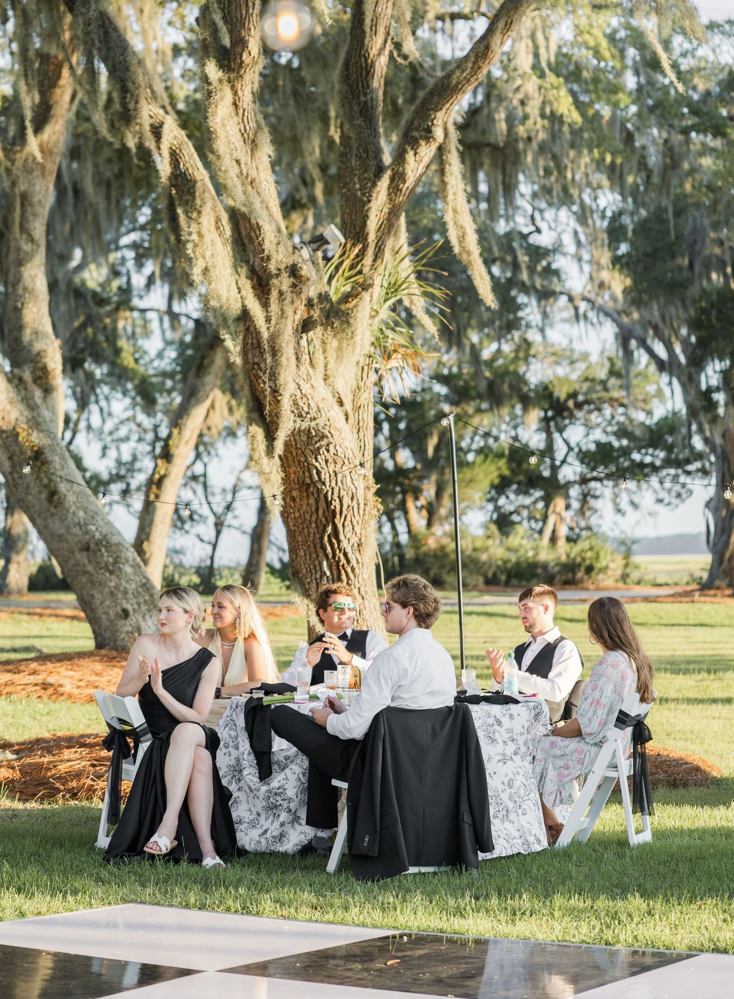 Wedding guests in black formal attire dining at outdoor reception table beneath Spanish moss-draped oak tree.