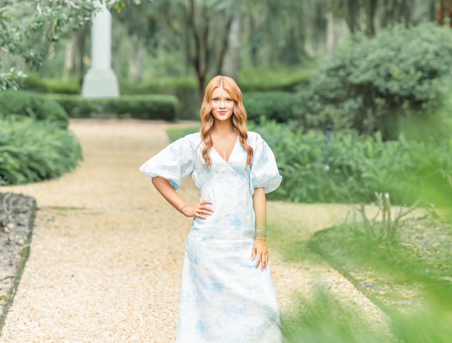 Someone in a white dress poses elegantly in a lush summer garden setting with gravel paths and green foliage.