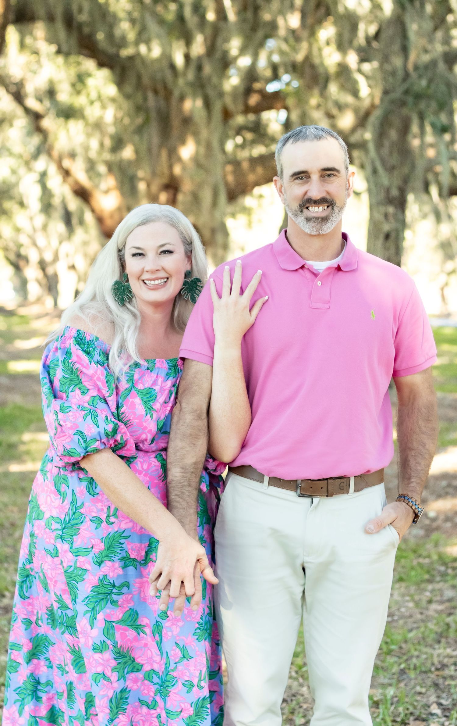 Couple smiles at camera in coordinated pink outfits against tree-lined background.