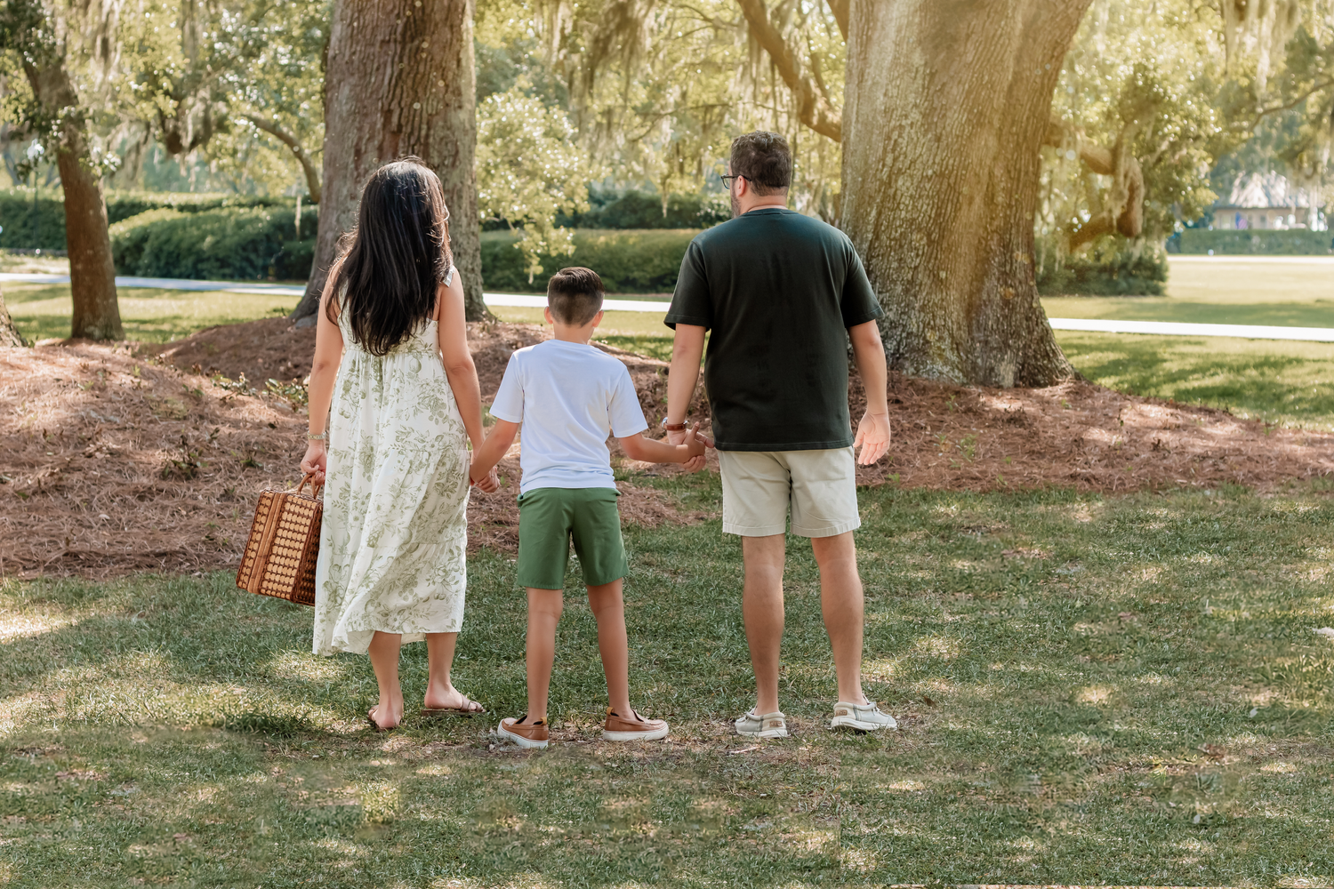 A family wearing casual attire stands together on a grassy area under large moss-draped oak trees in a park.