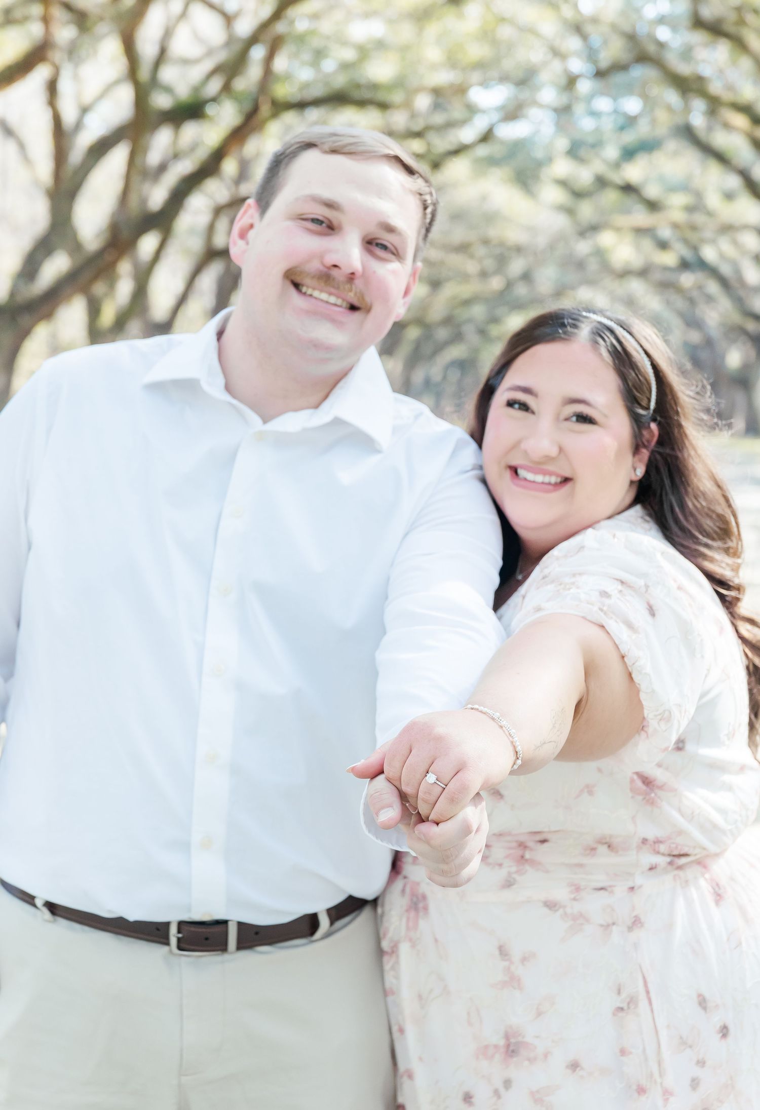 A couple smiling at the camera in a romantic outdoor photoshoot under an oak tree canopy.