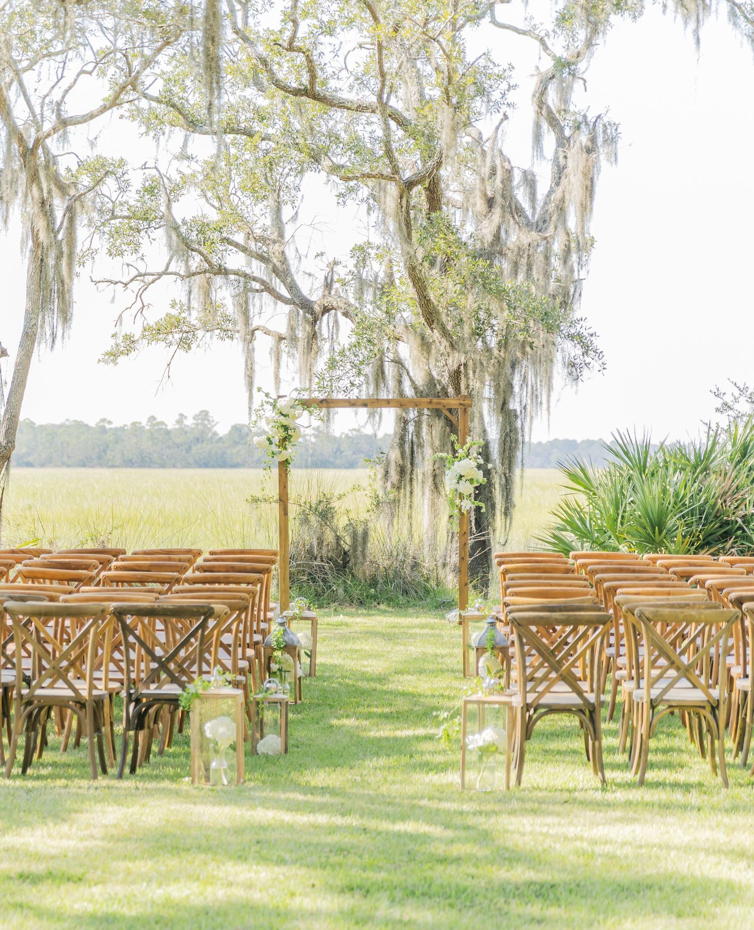 Rustic wooden chairs arranged for an outdoor wedding ceremony under Spanish moss-draped oak trees.