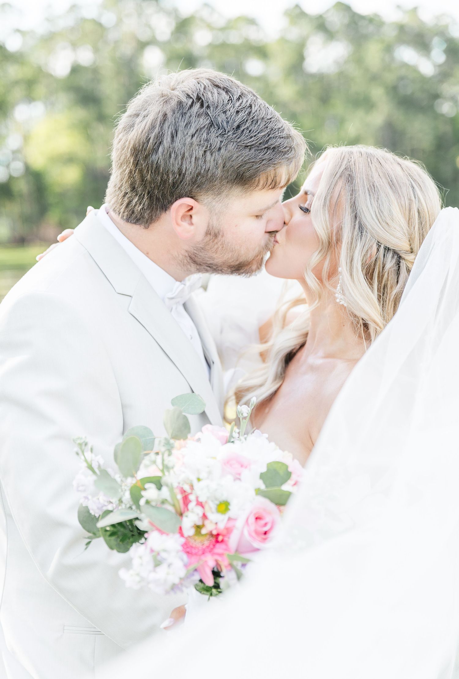 Light and airy wedding photo of couple's romantic kiss with soft pink floral bouquet and flowing veil.